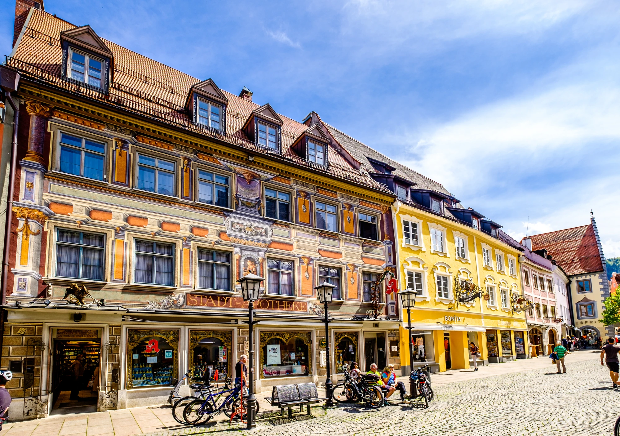Das Bild zeigt eine malerische Straßenszene in Füssen, geprägt von historischen Gebäuden mit kunstvollen Fassaden, die in hellen Farbtönen erstrahlen. Die Stadt ist bekannt für ihre gut erhaltene Altstadt, die von charmanten Geschäften und Cafés gesäumt wird. Im Vordergrund sind mehrere Fahrräder abgestellt, während Passanten die Straße entlang flanieren. Die Architektur spiegelt die Tradition und den Charakter dieser bayerischen Stadt wider, während das schöne Wetter und der klare Himmel eine einladende Atmosphäre schaffen.