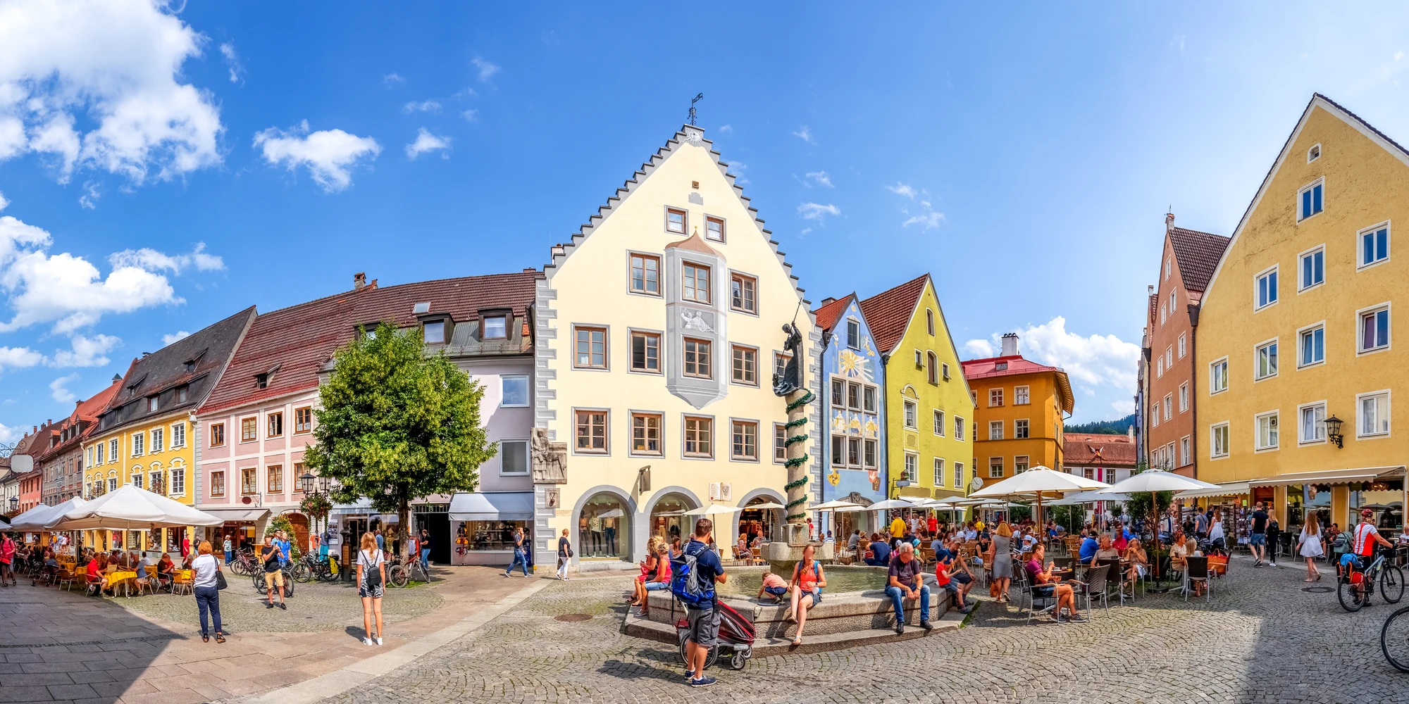 Das Bild zeigt den lebendigen Marktplatz in Füssen, umgeben von historischen Gebäuden mit bunten Fassaden, die typisch für die Altstadt der bayerischen Stadt sind. Auf dem Platz sind Menschen zu sehen, die in den Cafés entspannen, während Passanten durch die malerische Umgebung schlendern. Die einladende Atmosphäre wird durch das klare Wetter und die modernen Terrassenmöbel unter den Sonnenschirmen verstärkt. Im Hintergrund erheben sich die charakteristischen Dächer der traditionellen Häuser und eine markante Architektur ist ebenfalls gut erkennbar.