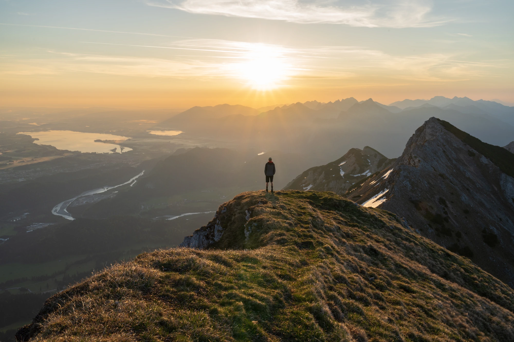 Das Bild zeigt einen atemberaubenden Blick auf Füssen und die umliegenden Berge, aufgenommen während des Sonnenuntergangs. Ein Wanderer steht am Rand eines Gipfels und genießt die Aussicht auf das weite Tal, die schneebedeckten Bergspitzen und den klaren Himmel. Die goldenen Lichtstrahlen der untergehenden Sonne tauchen die Landschaft in warmes Licht, das mit der grünen Wiese und den sanften Hügeln harmoniert. Der idyllische Moment zeigt die ruhige Schönheit der Natur rund um Füssen.
