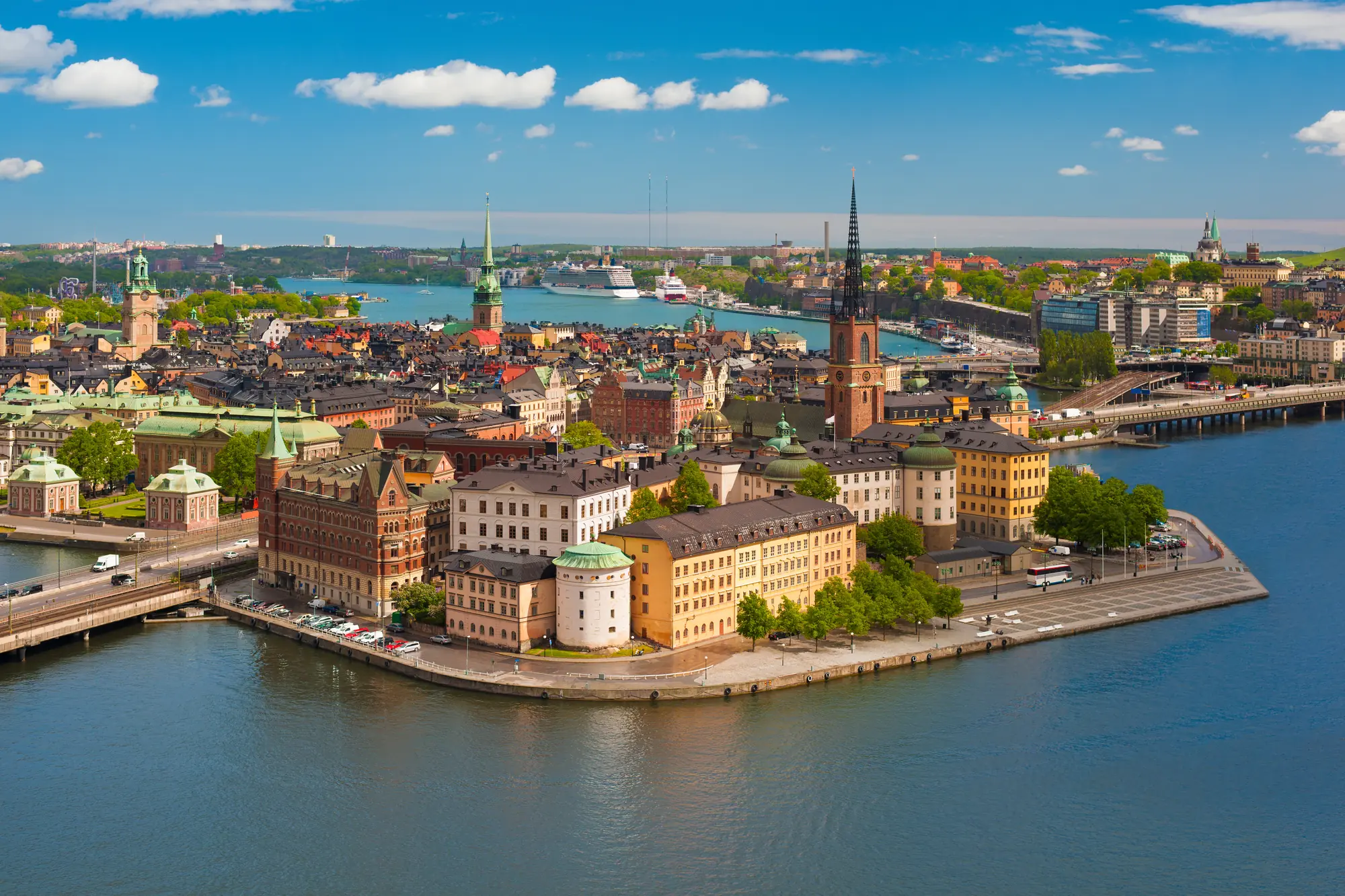 Luftaufnahme von Gamla Stan, der historischen Altstadt Stockholms, umgeben von Wasser und geprägt von malerischen, farbenfrohen Gebäuden. Im Zentrum ragt die markante Riddarholmskyrkan mit ihrem filigranen Turm empor. Im Hintergrund erstreckt sich die moderne Stadt mit Wasserwegen, Brücken und Fähren, die das Stadtbild prägen.
