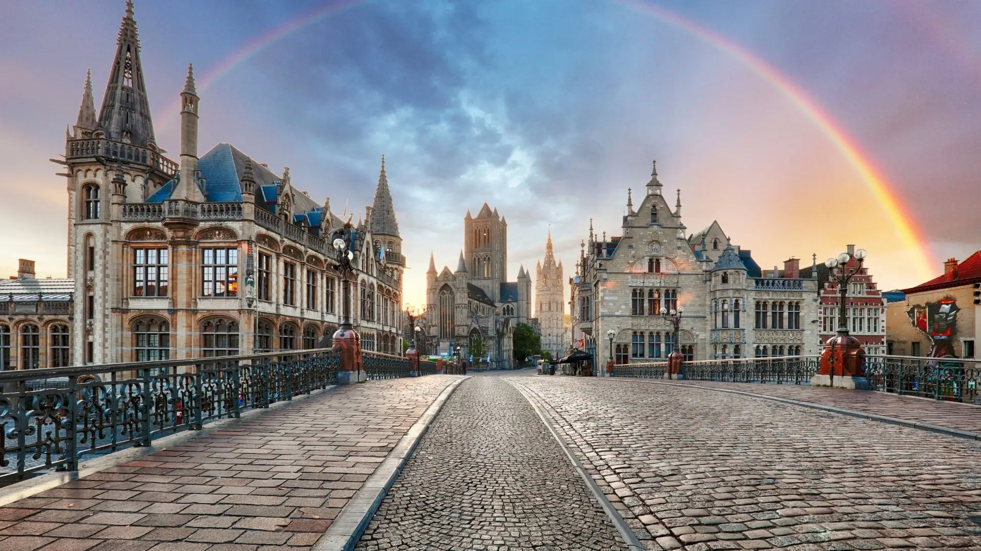 Blick über eine gepflasterte Brücke in Gent mit historischen Gebäuden und einem Regenbogen am Himmel.