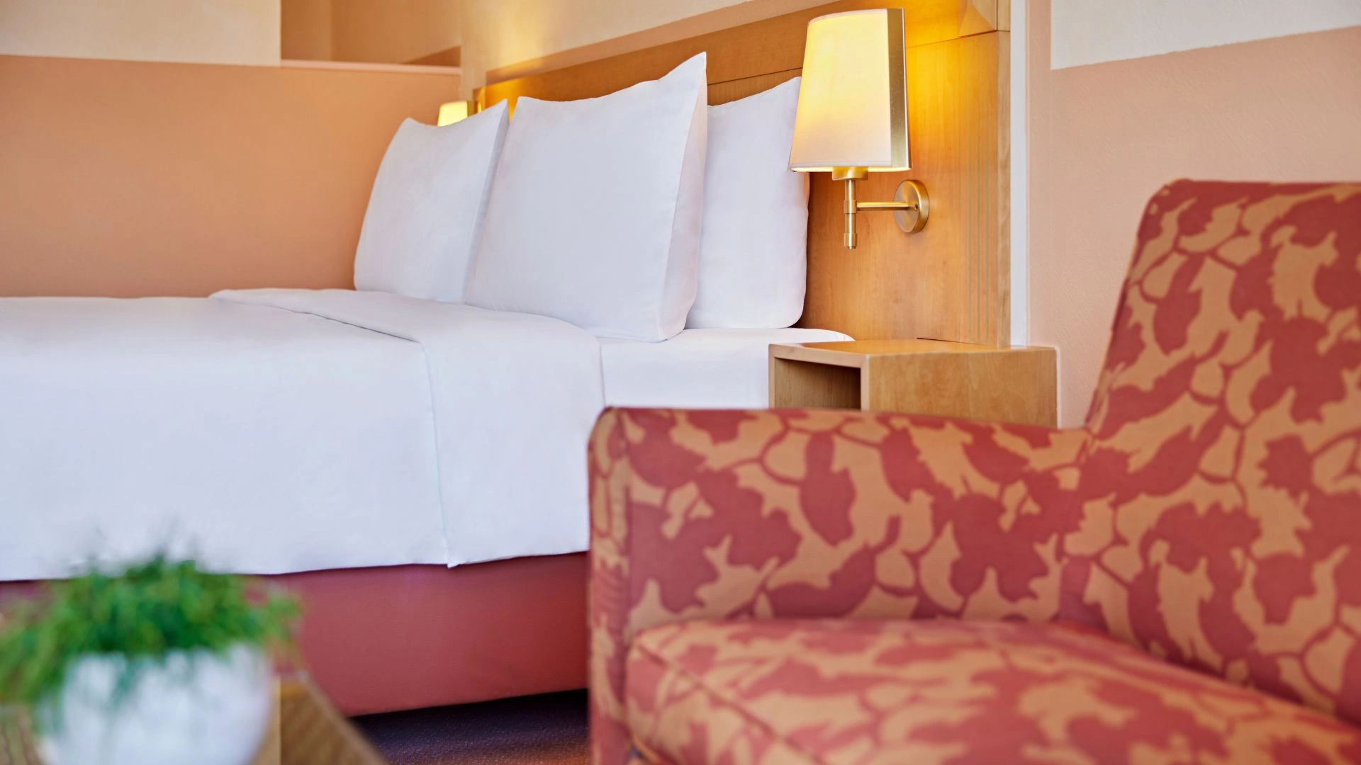 Hotelzimmer mit großem Bett, weißen Kissen, Holzwand mit Wandlampe und gemustertem Sessel im Vordergrund