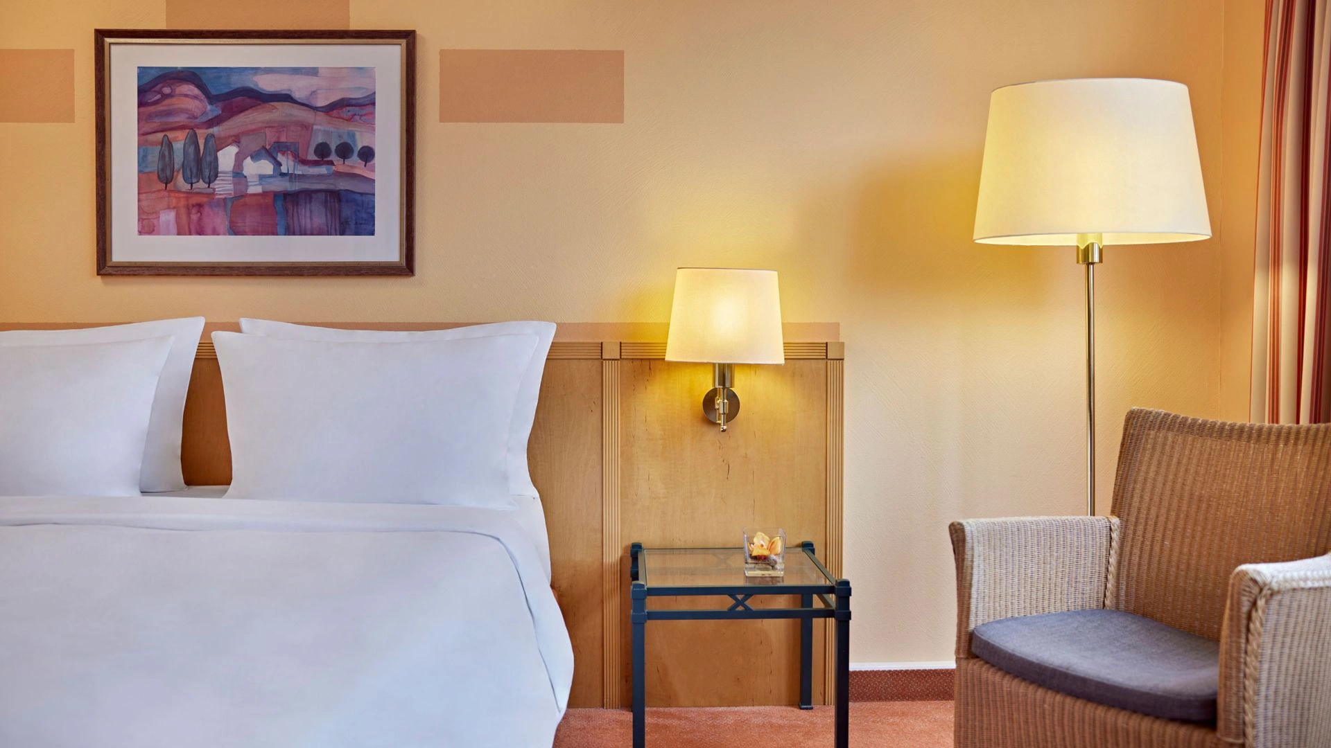Hotelzimmer mit Doppelbett, Nachttisch mit Lampe, Sessel und Stehlampe vor beiger Wand mit farbiger Landschaftsmalerei