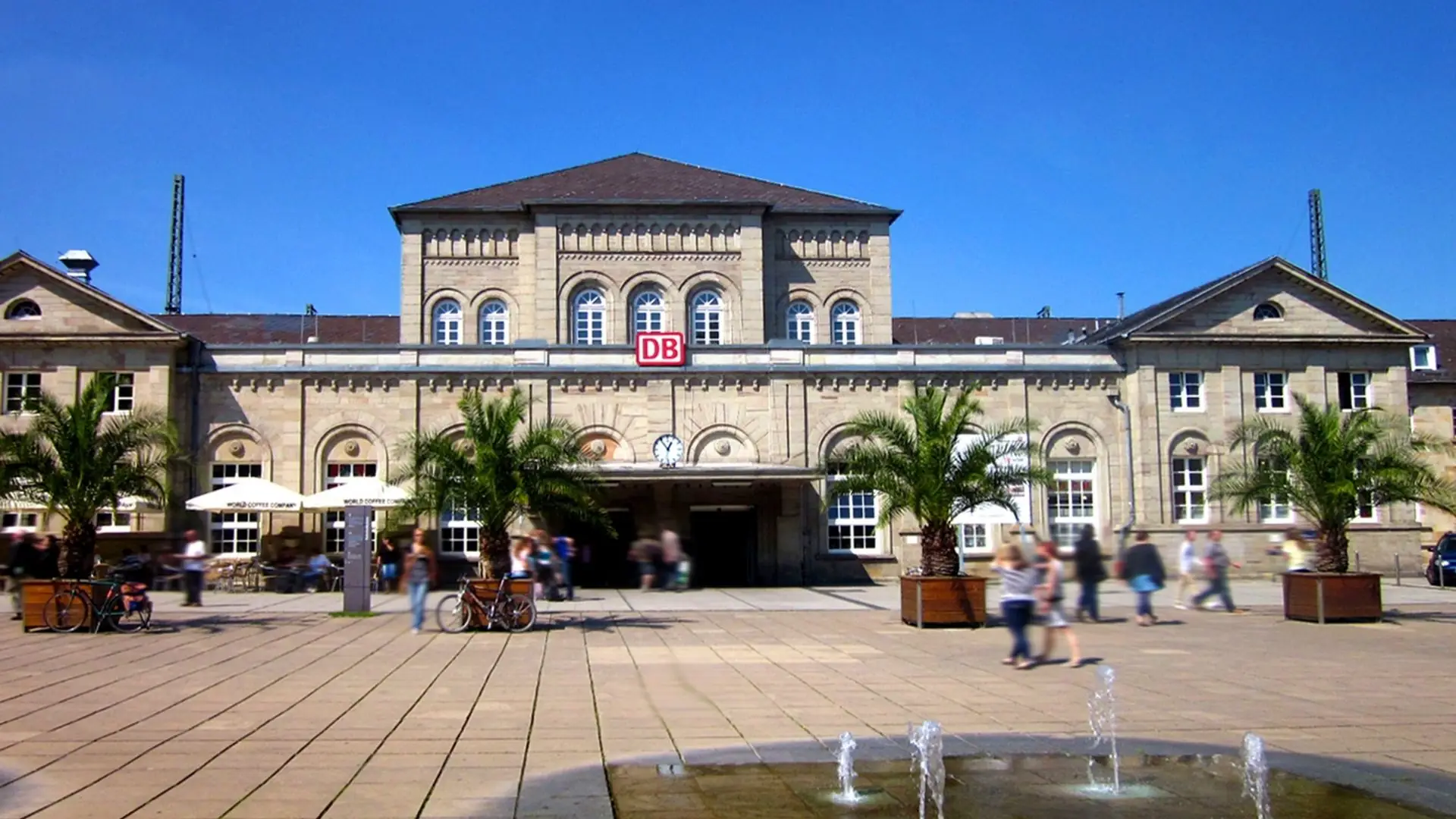 Hauptgebäude des Göttinger Hauptbahnhofs mit der DB-Logo über dem Eingang. Vor dem Gebäude stehen Palmen und ein Brunnen, viele Menschen sind auf dem Platz.