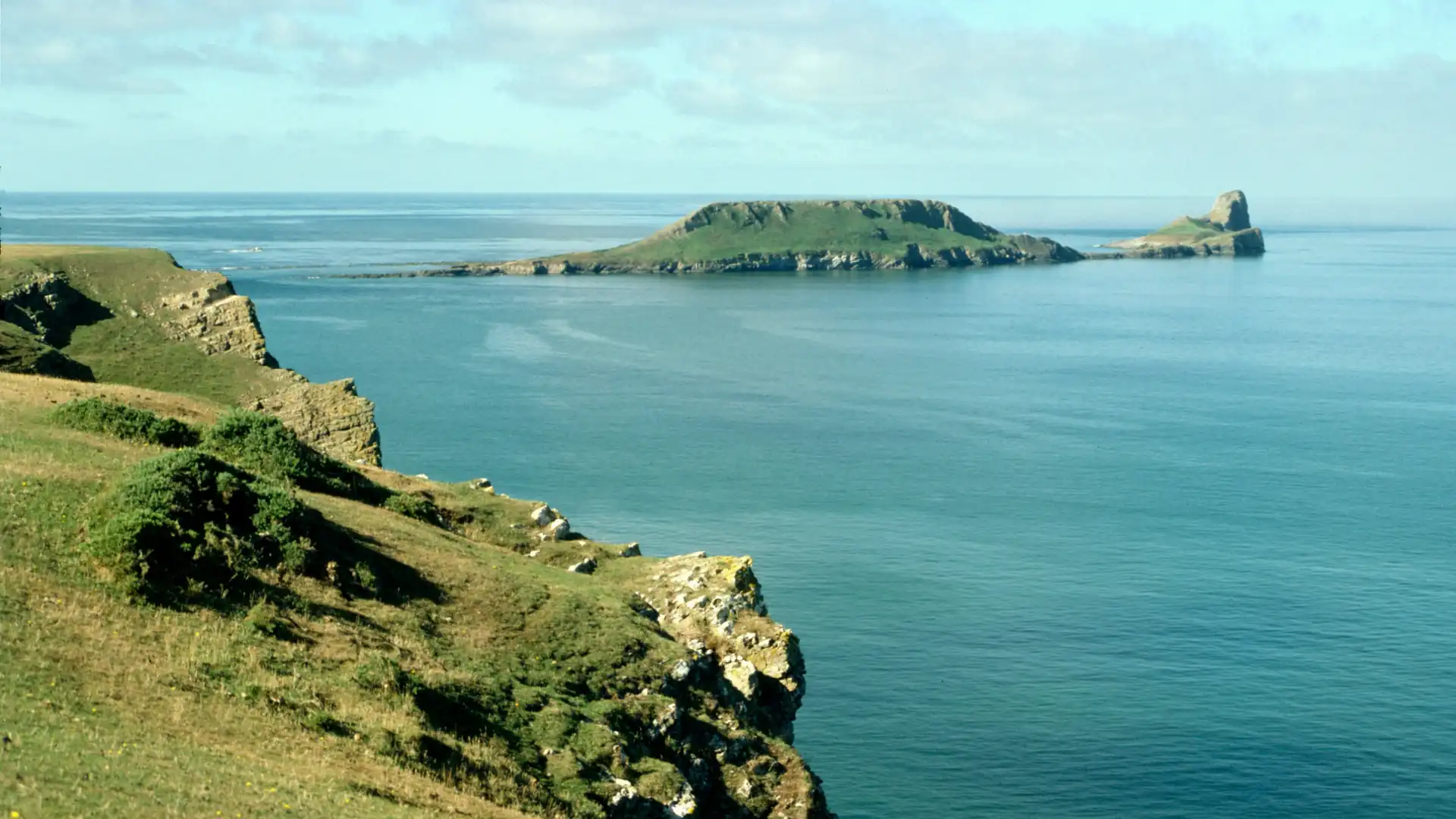 Grüne Klippen der Gower Halbinsel mit Büschen am Meer, im Hintergrund eine flache Insel unter bewölktem Himmel