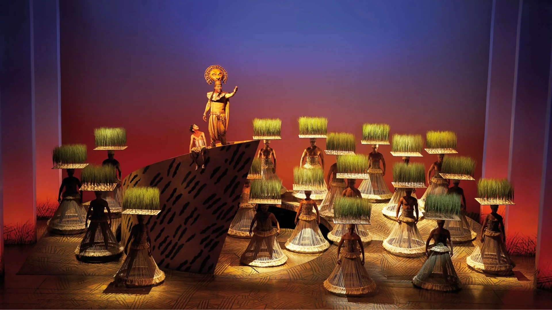 Eine lebhafte Bühnenszene mit Darstellern in traditioneller Kleidung, die vor einem dramatischen Sonnenuntergang mit Gras balancieren.