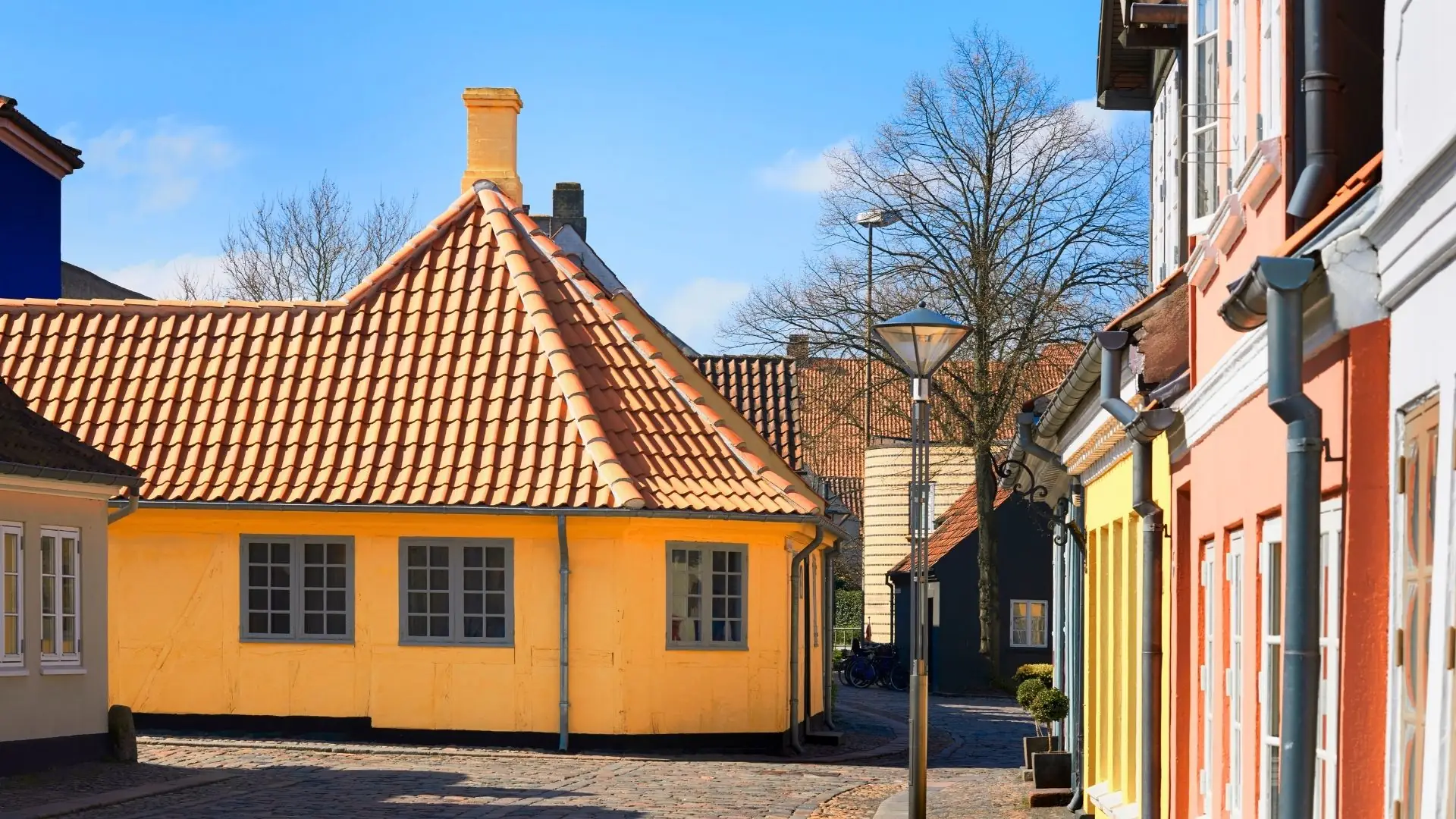 Gelbes, historisches Fachwerkhaus mit rotem Ziegeldach und mehreren Fenstern, umgeben von weiteren Gebäuden und Bäumen im Hintergrund.