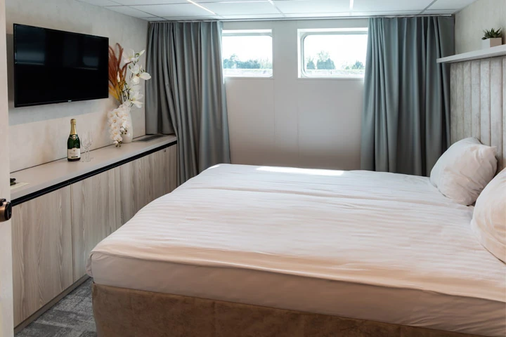 Doppelbett mit weißen Kissen und Bettwäsche in einem Zimmer mit zwei kleinen Fenstern, grauen Vorhängen, Wandfernseher und Flasche auf Sideboard