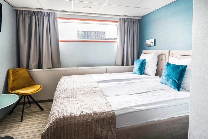 Kabine mit zwei Einzelbetten, blauen Kissen, grauer Wand, gelbem Stuhl und Fenster mit grauen Vorhängen