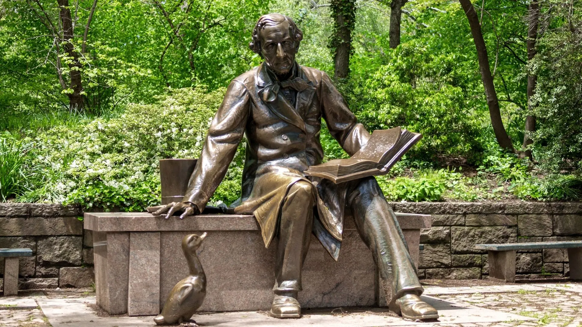 Bronzestatue von Hans Christian Andersen sitzend auf einer Steinbank, ein aufgeschlagenes Buch auf dem Schoß, umgeben von grünem Laub.