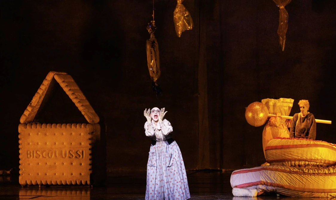 Theaterszene mit einer Frau in historischer Kleidung, die überrascht oder erschrocken wirkt, vor einer großen Keksrequisite mit der Aufschrift 'BISCOTUSSI' und einem Mann neben einem übergroßen Musikinstrument.