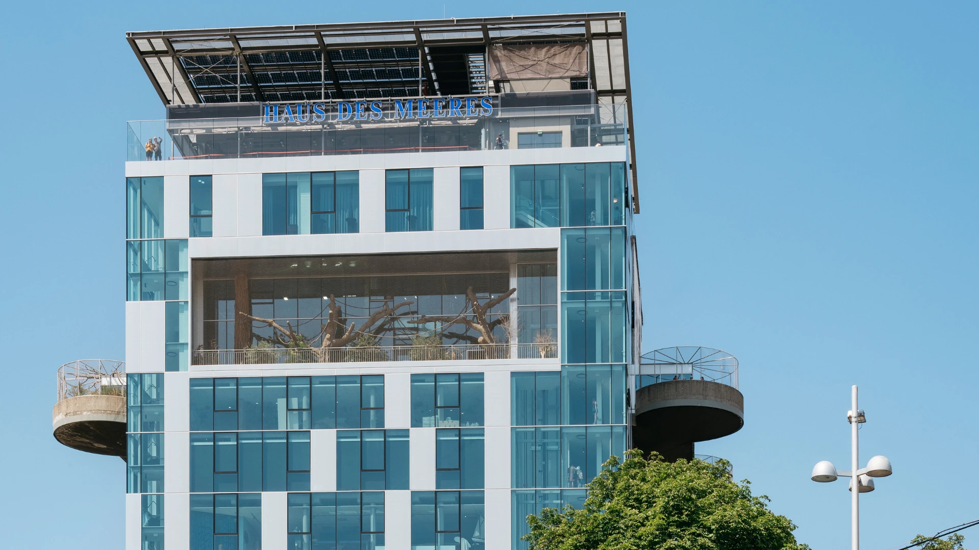Dieses Foto zeigt das Haus des Meeres in Wien, ein modernes Aquarium und Tropenhaus in einem ehemaligen Flakturm. Die markante Glasfassade reflektiert den Himmel, während im offenen Bereich eine Affenanlage zu sehen ist. Auf der Dachterrasse genießen Besucher den Panoramablick über die Stadt.