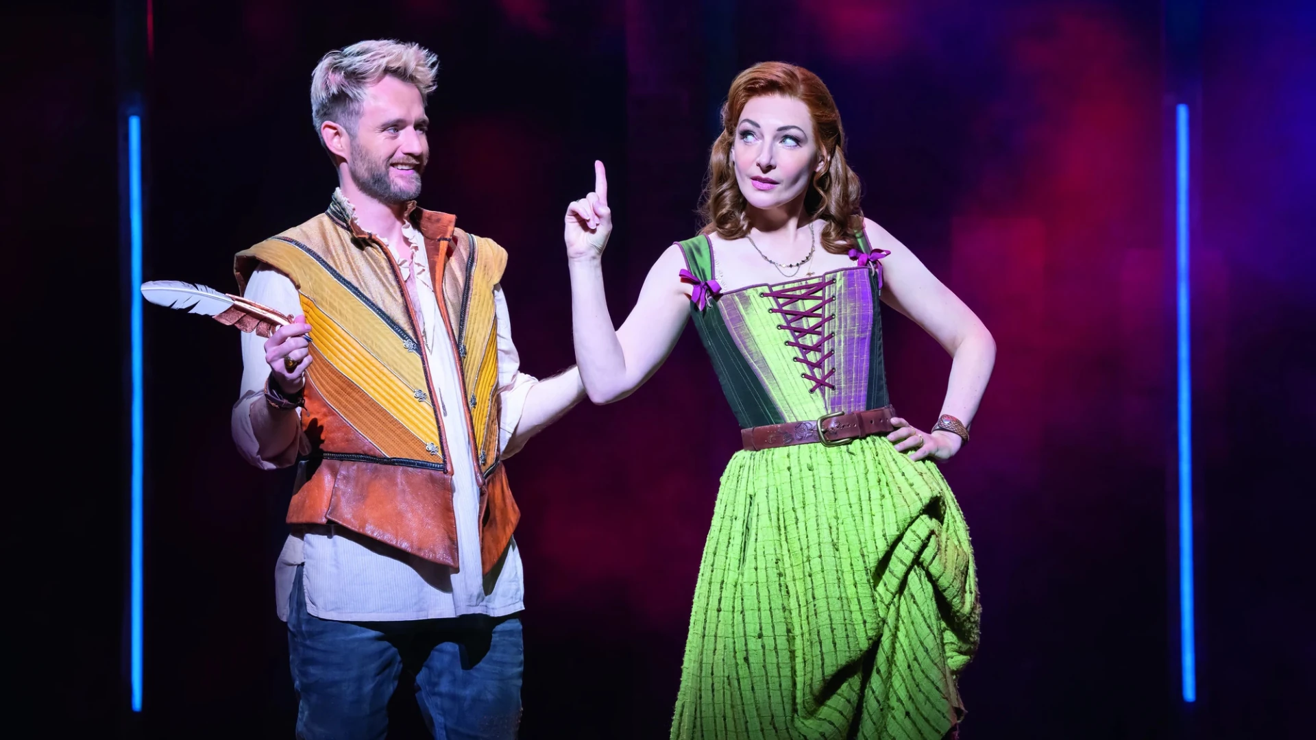 Zwei Darsteller auf der Bühne: einer in einer bunten Weste mit einer Feder in der Hand, der andere in einem leuchtend grünen Kleid, beide in dynamischen Posen.