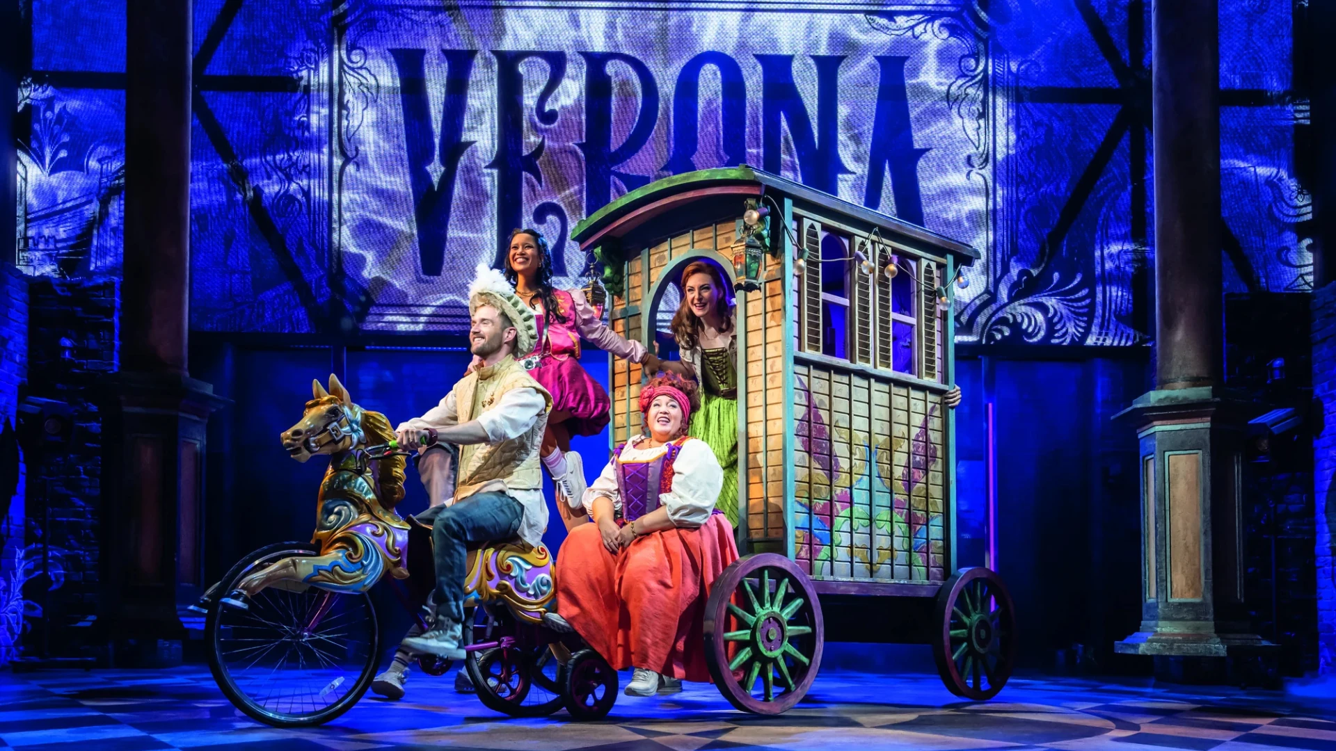 Eine lebendige Theaterszene mit Darstellern auf einem skurrilen Wagen und einer dekorativen Kulisse mit der Aufschrift „VERONA“.