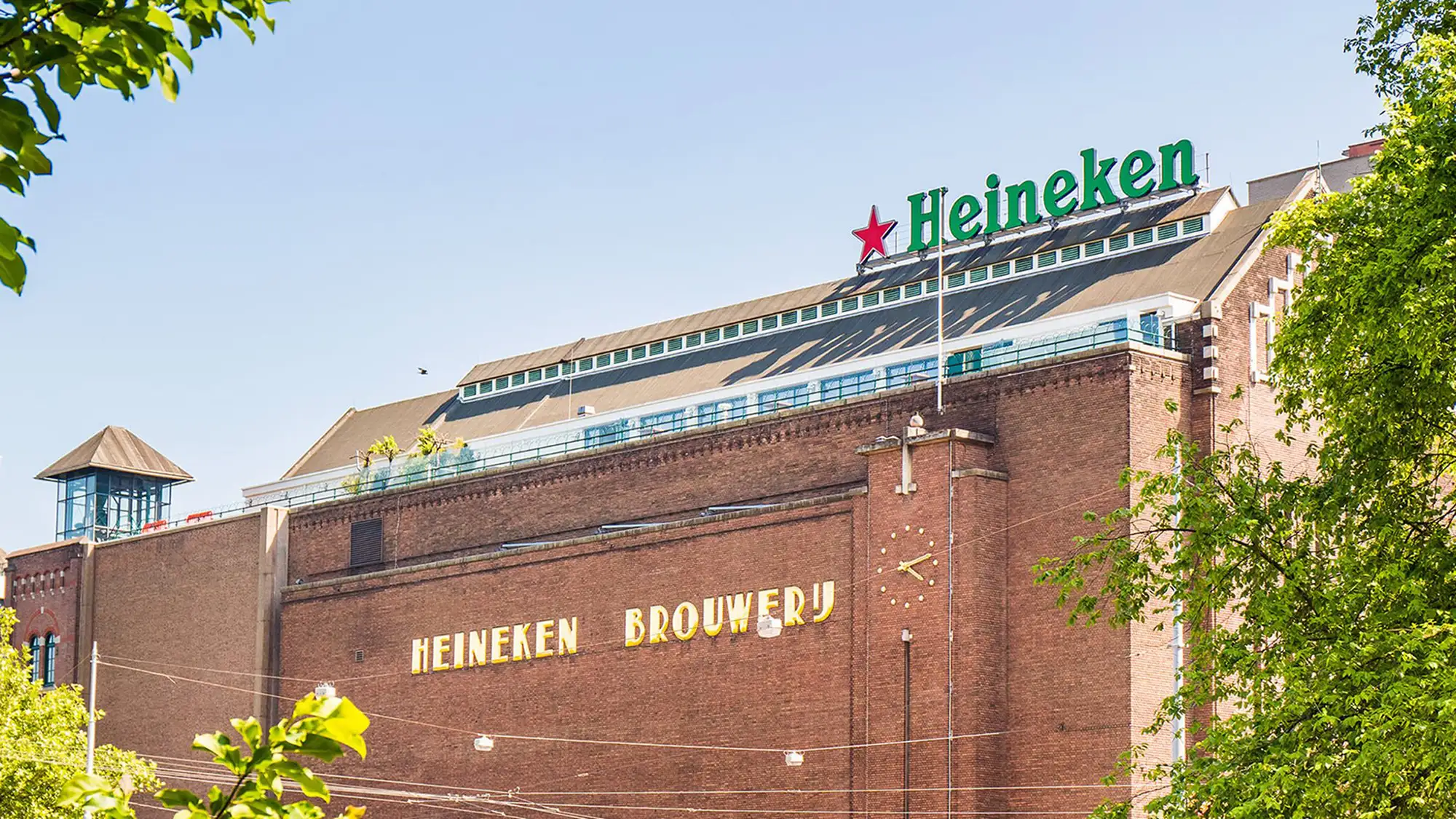 Das Bild zeigt die Außenansicht der historischen Heineken Brauerei in Amsterdam. Das Backsteingebäude wird von dem bekannten grünen Heineken-Schriftzug mit dem roten Stern gekrönt. Im Vordergrund rahmen grüne Bäume das Bild ein, und der klare Himmel verstärkt die Helligkeit der Szene. Das Gebäude symbolisiert die lange Geschichte der Brauerei und dient heute als Teil der Heineken Experience, einer beliebten Touristenattraktion.