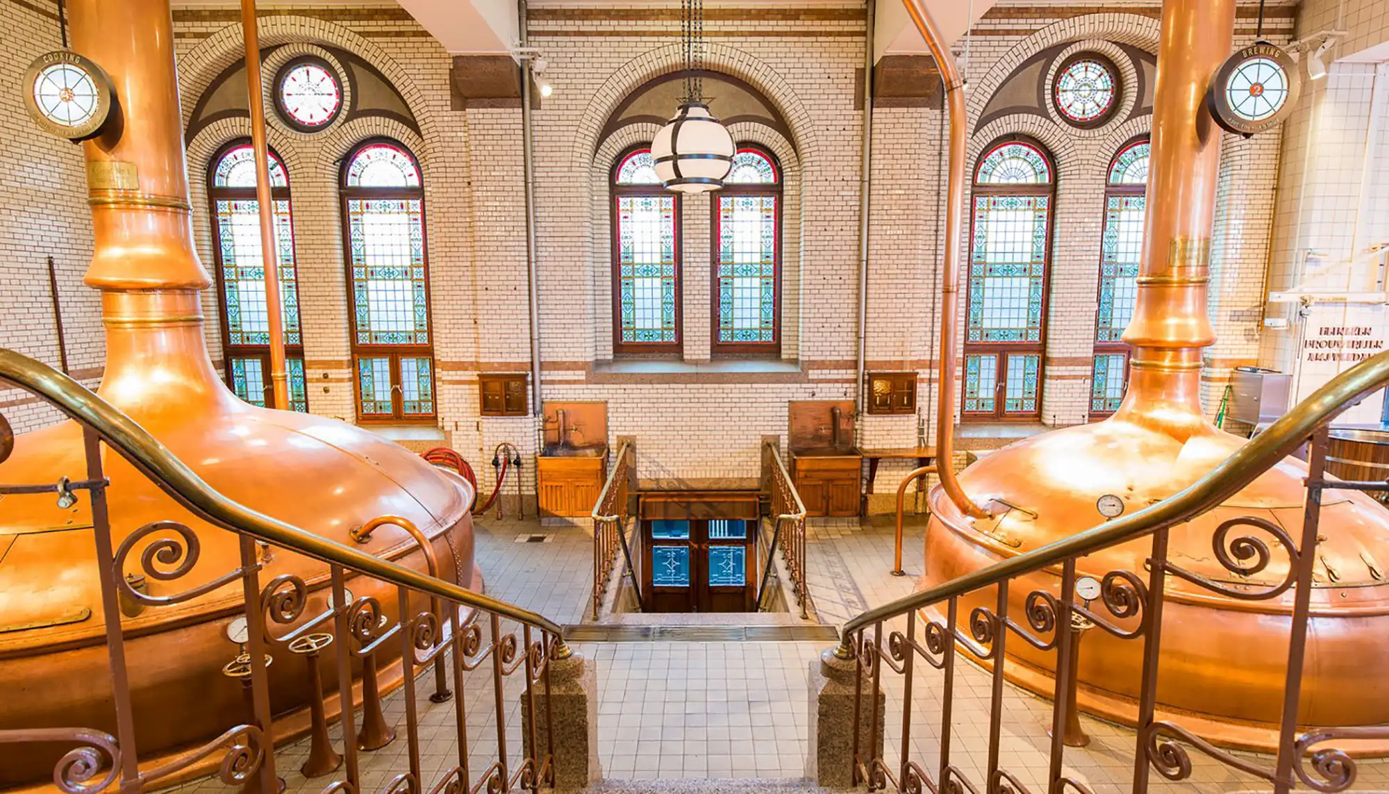 Das Bild zeigt eine Treppe im Heineken Museum in Amsterdam, die hinunter in den historischen Braukeller führt. Die Umgebung ist geprägt von großen, glänzenden Kupferkesseln, die traditionell für den Brauprozess verwendet wurden. Die hohen Fenster mit buntem Glas und die dekorativen Geländer verleihen dem Raum ein klassisches und stilvolles Ambiente, das die Geschichte und Tradition der Heineken-Brauerei widerspiegelt.