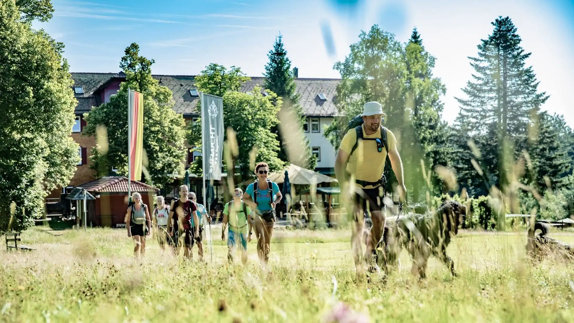 Gruppe von Wanderern mit Rucksack und Hund auf einer Wiese vor dem Natur- & Sporthotel Zuflucht mit Fahnen und Bäumen im Hintergrund