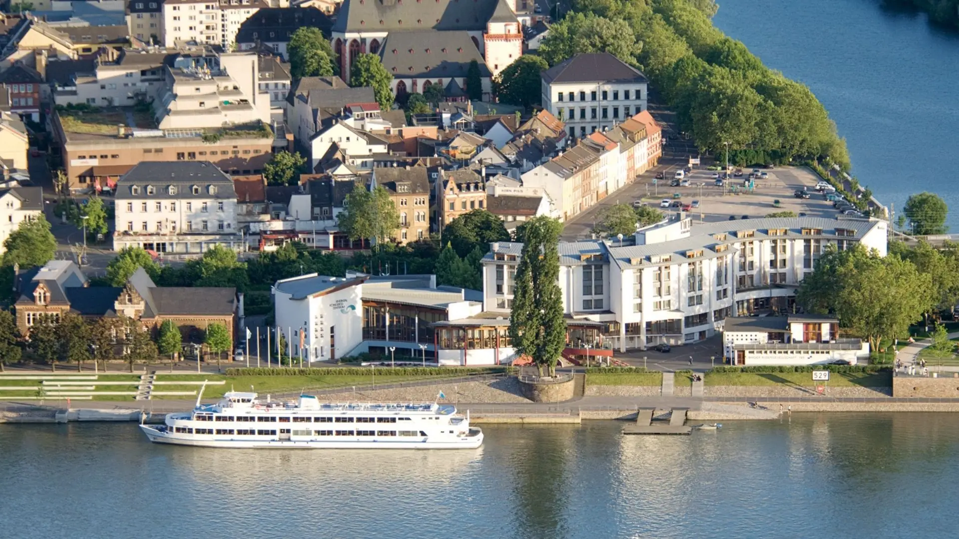 Luftaufnahme vom Hotel NH Bingen mit Flussufer, mehreren Gebäuden und einem weißen Ausflugsschiff am Anleger