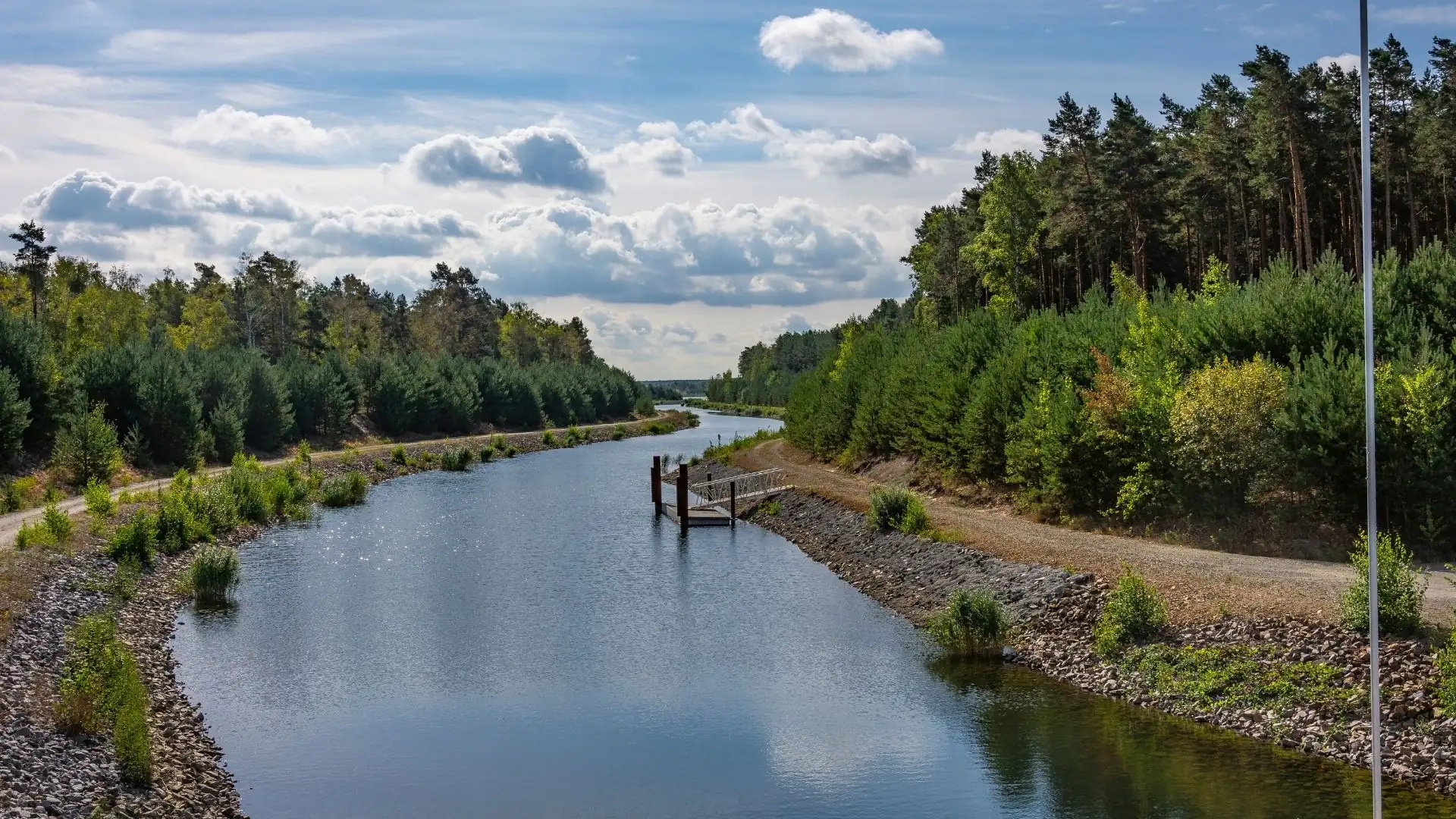 Blick auf einen ruhigen Kanal im Lausitzer Seenland, umgeben von bewaldeten Uferbereichen und einem Kiesweg unter einem bewölkten Himmel