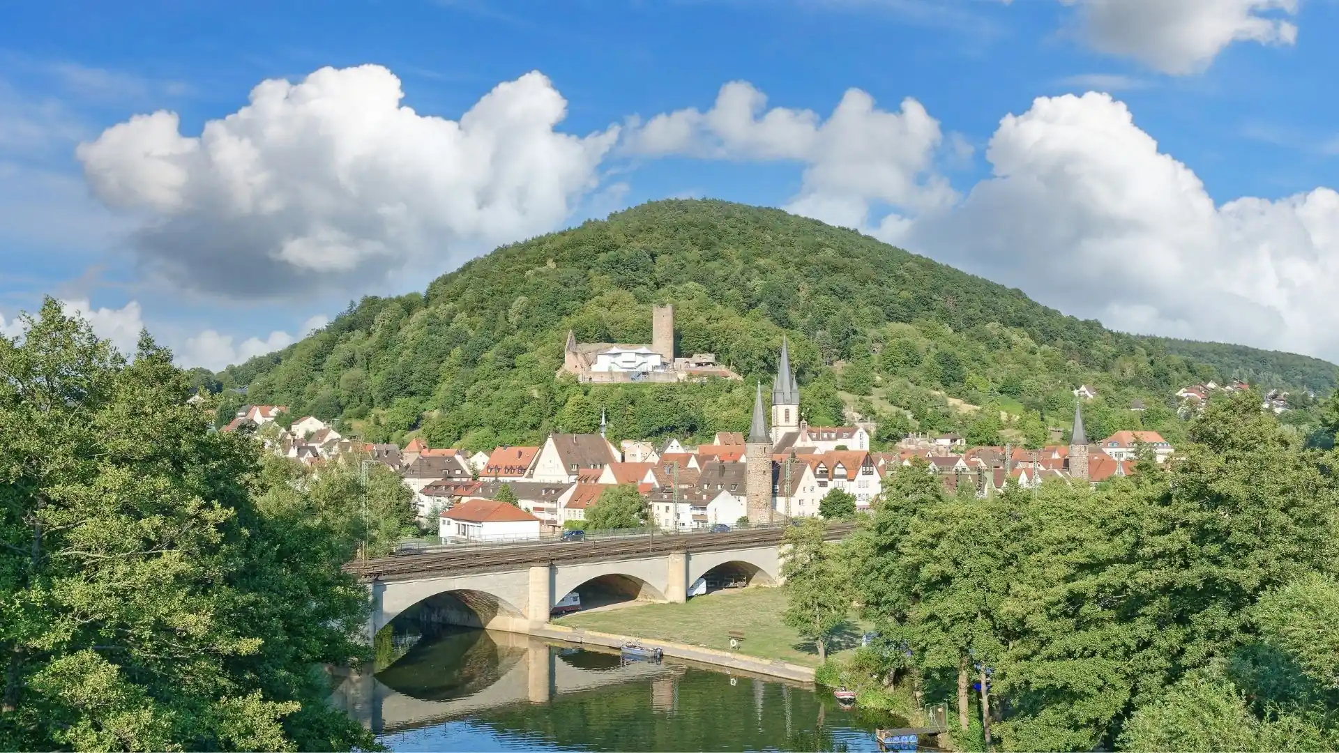 Blick auf Gemünden am Main mit Brücke über den Fluss, historischen Gebäuden und bewaldetem Hügel im Hintergrund unter blauem Himmel mit Wolken