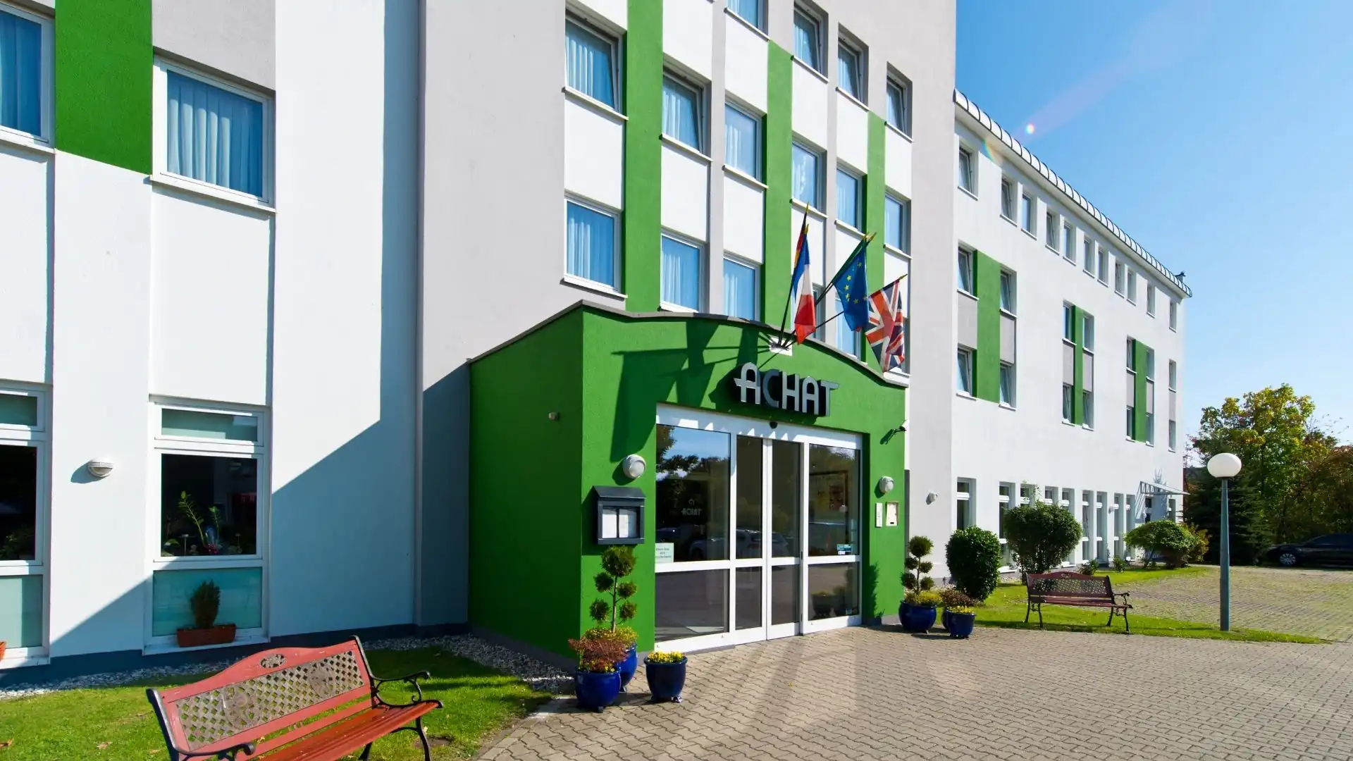 Dreigeschossiges Hotelgebäude mit weißer Fassade und grünen Fensterläden, Eingangsbereich mit grünem Vordach und Schriftzug 'ACHAT'