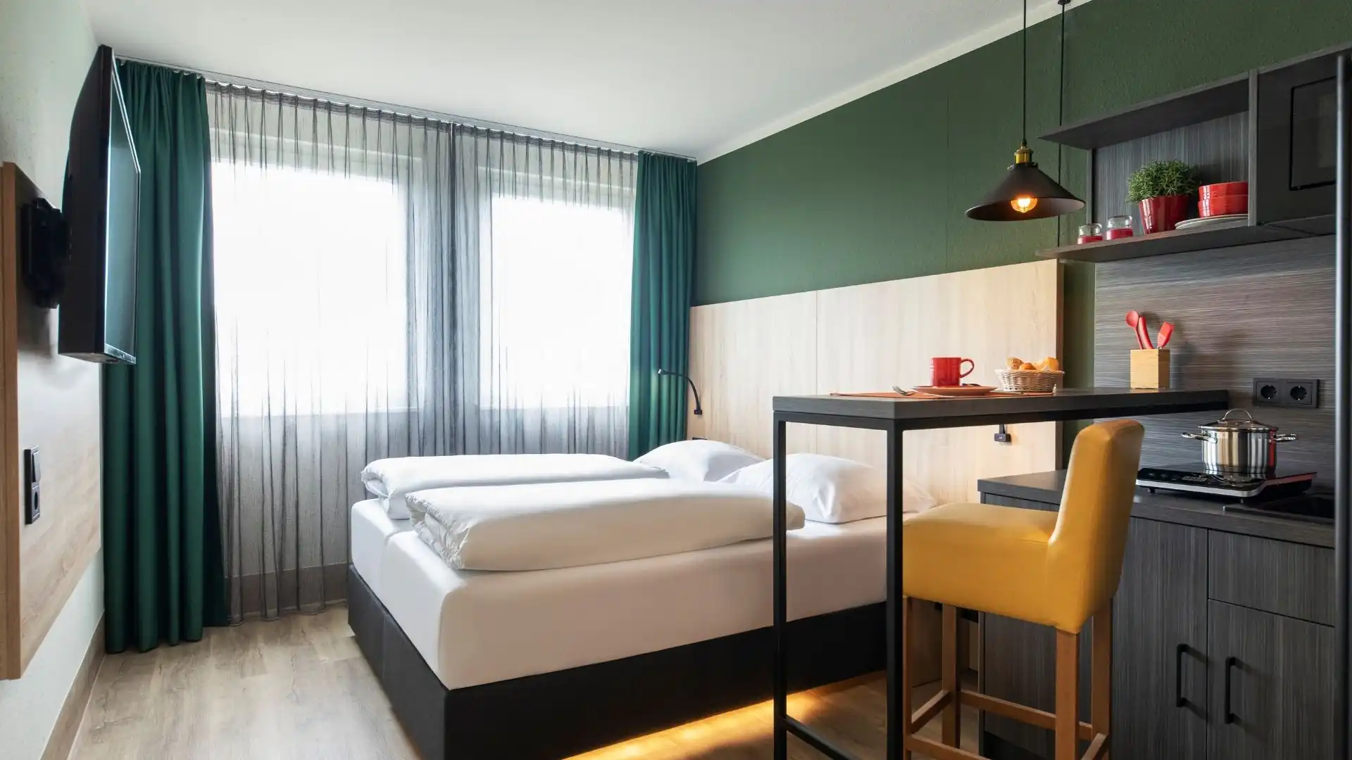 Hotelzimmer mit zwei Einzelbetten, Nachttischlampen, grünem Vorhang, Schreibtisch mit Wasserflaschen und Gläsern sowie einem an der Wand montierten Fernseher