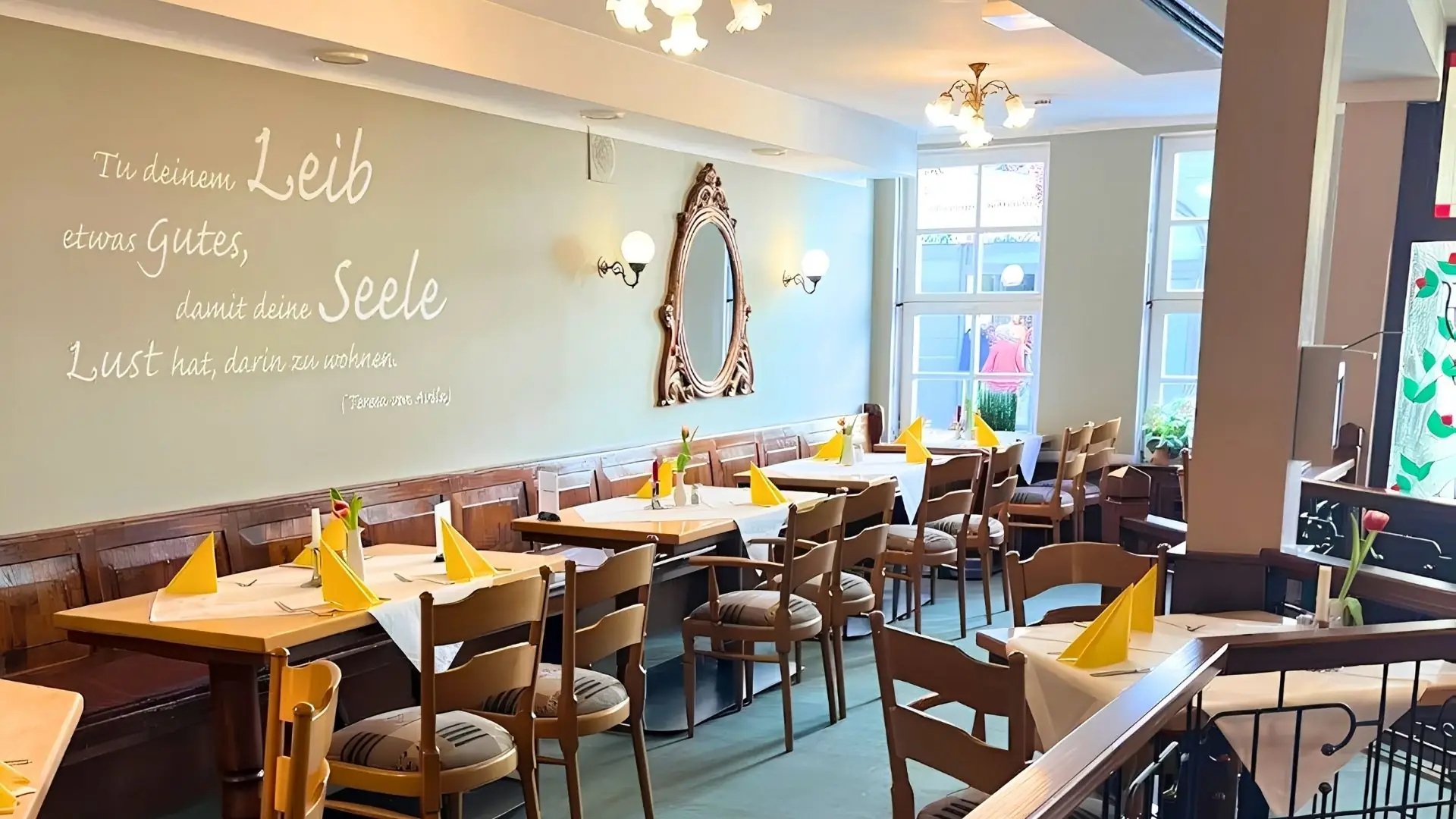 Restaurant mit Holztischen und -stühlen, gelben Servietten auf den Tischen, Wand mit dekorativem Spiegel und Schriftzug