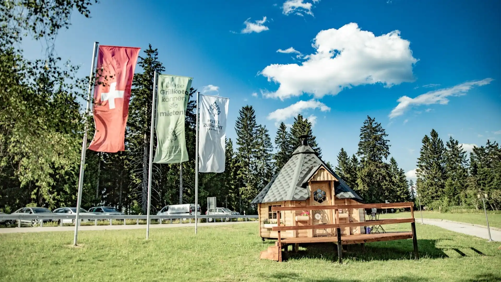 Kleines hölzernes Uhrhaus mit Spitzdach auf einer Wiese neben vier Fahnenmasten vor Nadelbäumen und blauem Himmel mit Wolken