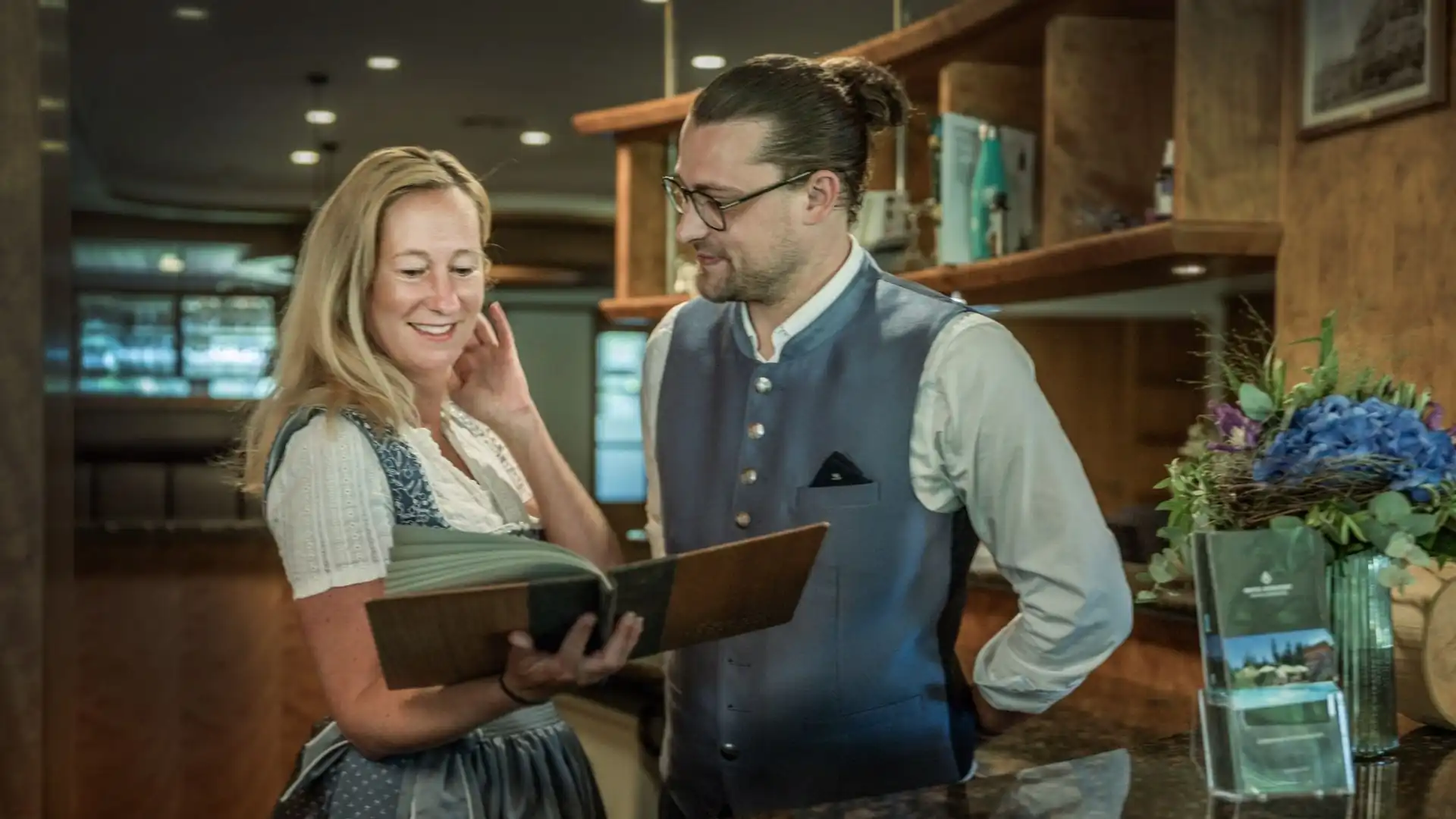 Zwei Personen in traditioneller Tracht stehen im Empfangsbereich und schauen gemeinsam in ein geöffnetes Buch