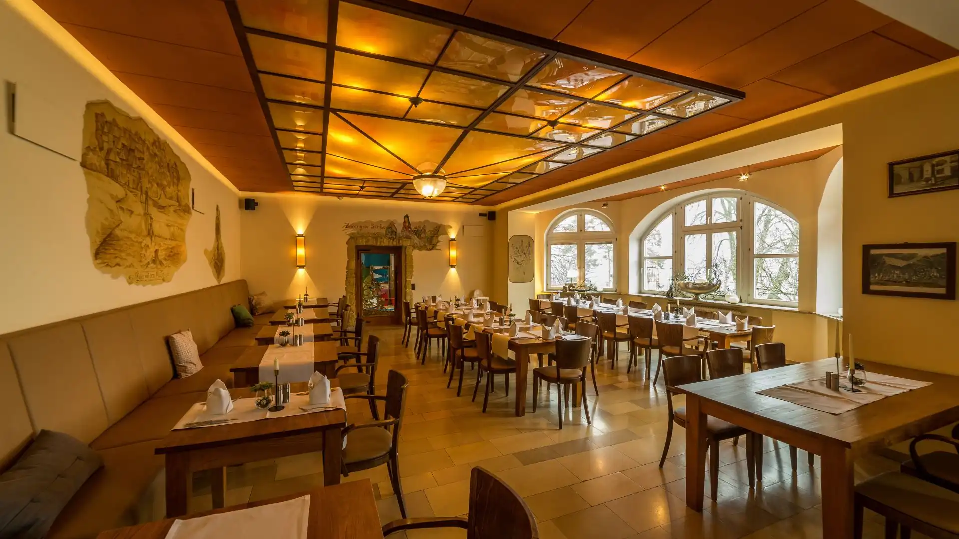 Restaurant mit Holztischen und Stühlen, langen Sitzbänken an der Wand, großen Fenstern und warm beleuchtetem Deckenraster im Hotel Franziskushöhe