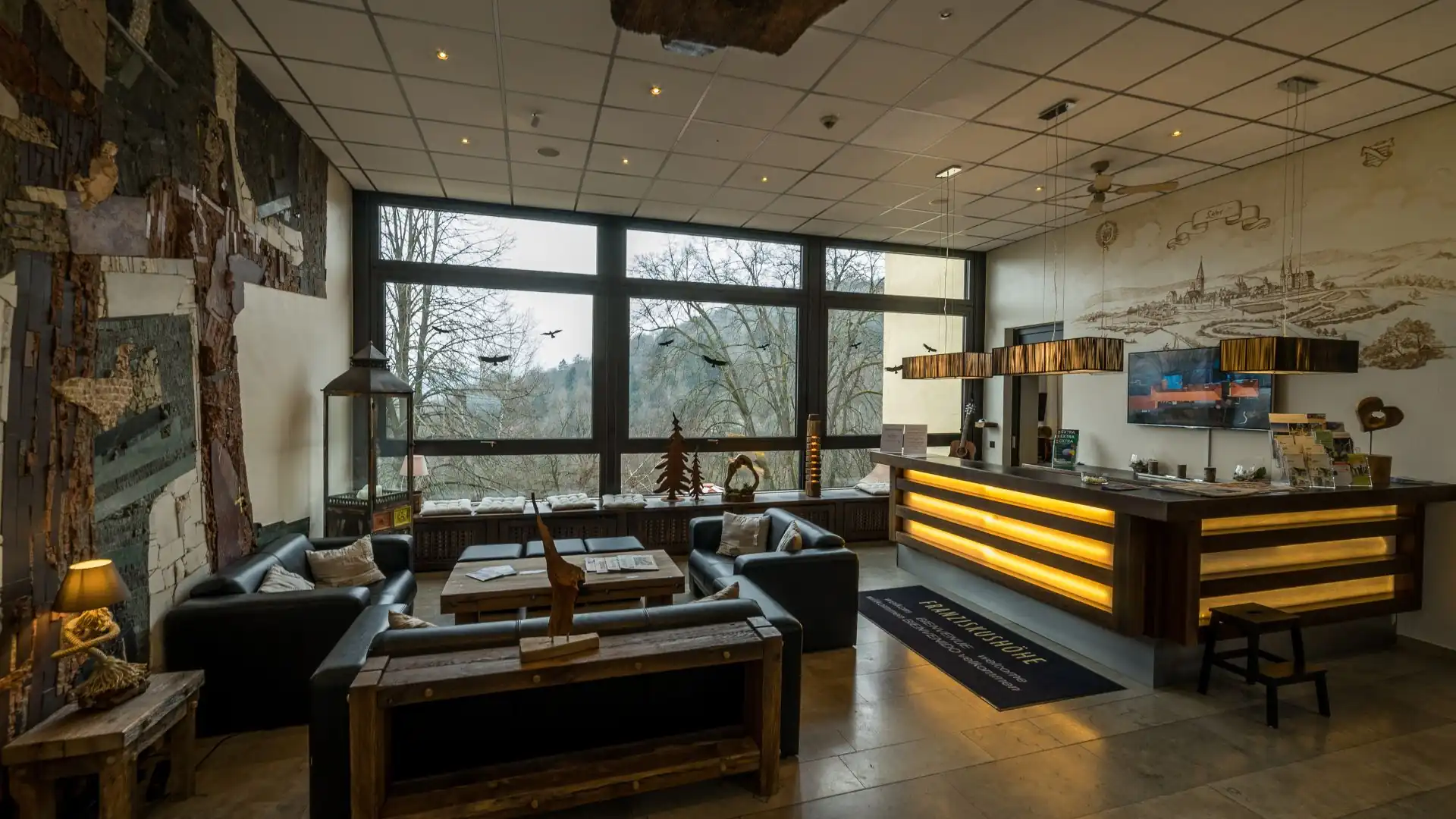 Hotelrezeption mit beleuchtetem Empfangstresen aus Holz, Sitzgruppe mit Sofas und Holztischen vor groĂen Fenstern mit Blick auf BĂ€ume und Landschaft.