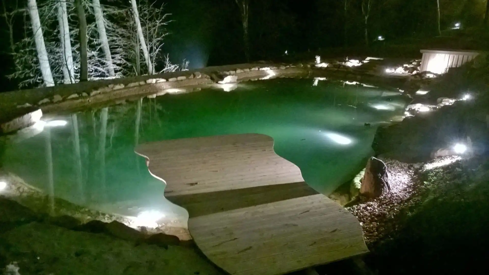 Naturnaher Schwimmteich mit Holzsteg und Beleuchtung bei Nacht umgeben von BÀumen und Felsen im Hotel Franziskushöhe