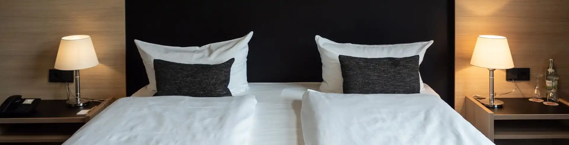 Doppelbett mit weißen Bettdecken und schwarzen Kissen vor einer holzvertäfelten Wand mit zwei Nachttischen und je einer Lampe