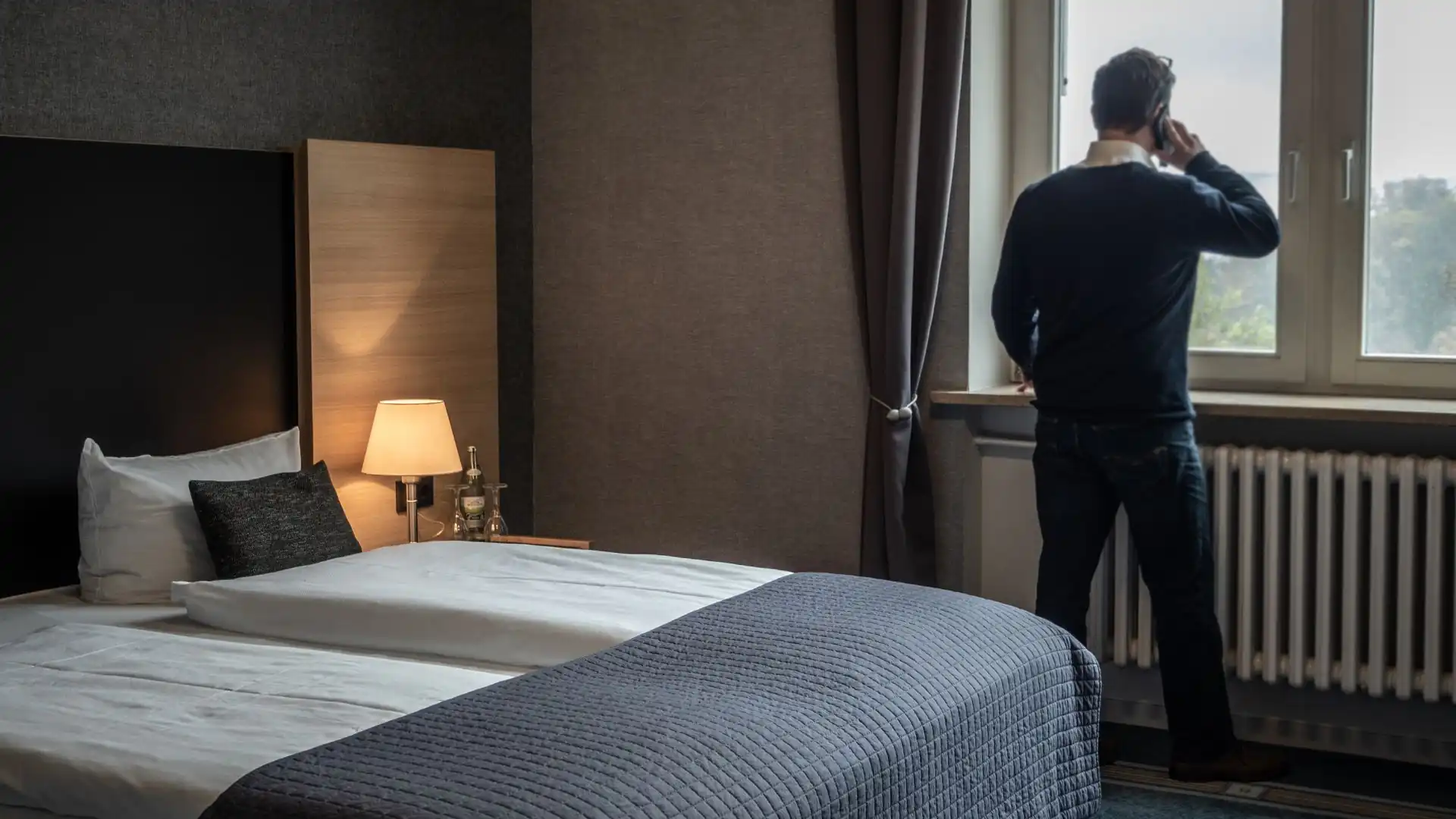 Mann steht am Fenster eines Hotelzimmers und telefoniert neben einem Bett mit weißer Bettwäsche und grauer Decke
