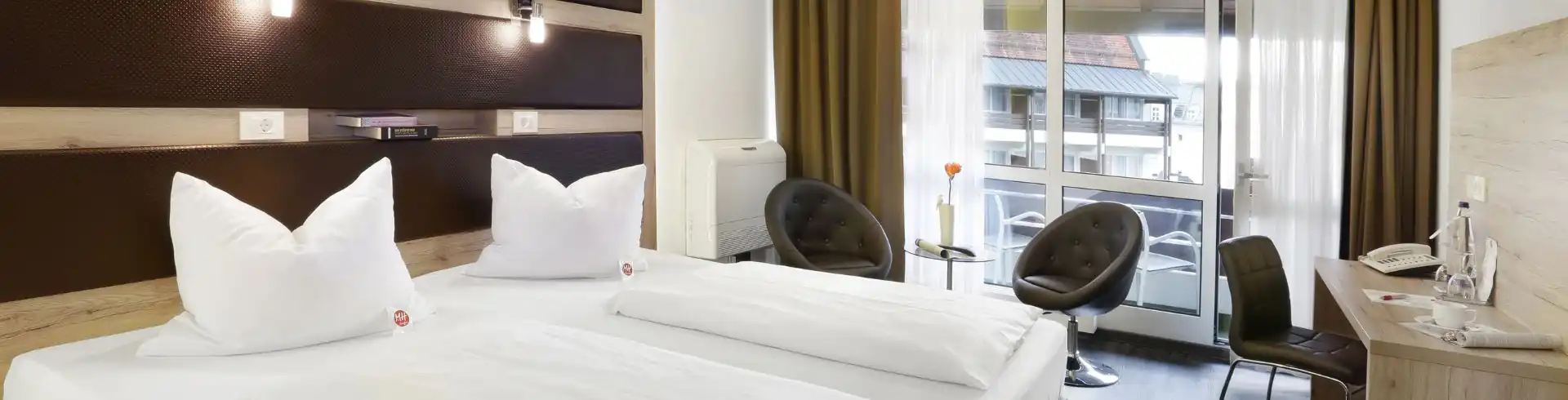 Hotelzimmer Banner mit zwei Einzelbetten, zwei Sesseln vor bodentiefem Fenster mit Balkon, Schreibtisch mit Stuhl und moderner Beleuchtung
