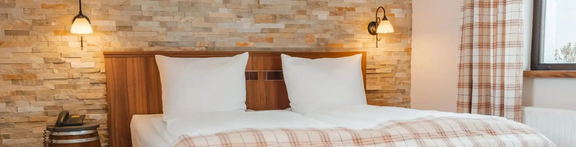 Doppelbett mit weißen Kissen vor einer Wand mit Natursteinverkleidung und zwei Wandlampen