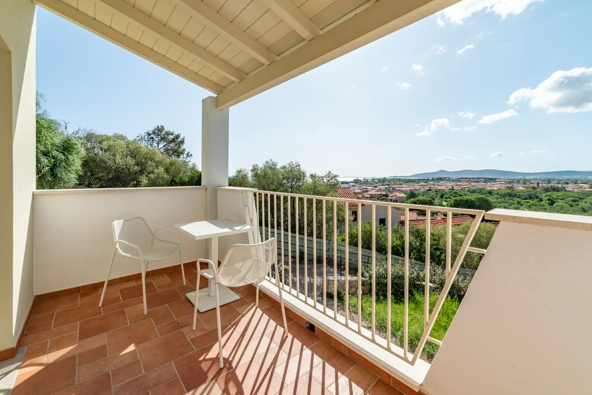 Balkonblick aus einem Zimmer des im Hotels Piccolo in Porto Ottiolu mit zwei weißen Stühlen und kleinem Tisch, Blick auf grüne Landschaft und Stadt im Hintergrund