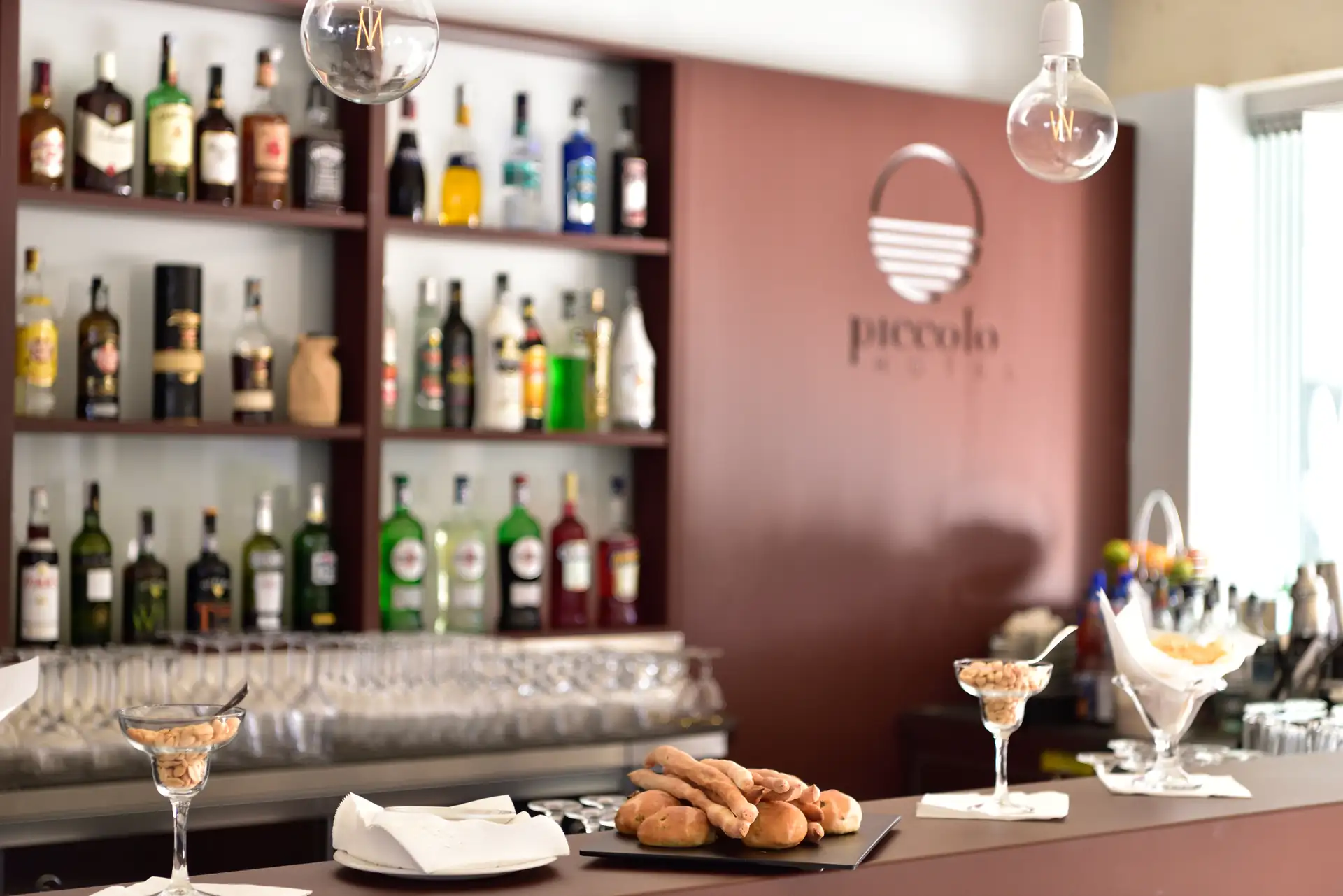 Bar im Hotel Piccolo in Porto Ottiolu mit Regalen voller Spirituosen, Tresen mit Snacks und Gläsern, Wand mit Logo 'piccolo'