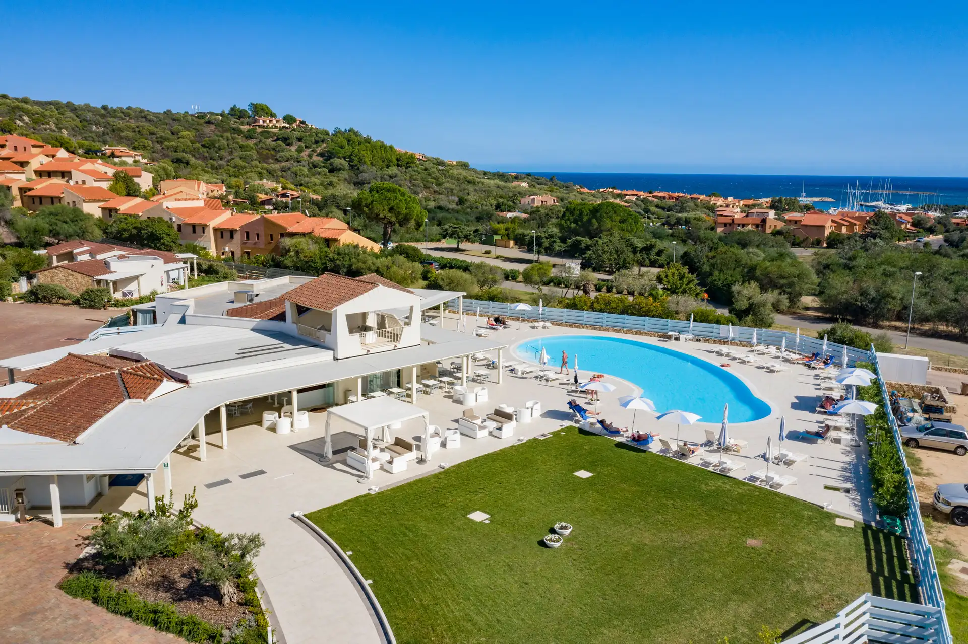 Großer Außenpool mit Liegestühlen und Sonnenschirmen neben dem Hotel Piccolo in Porto Ottiolu, umgeben von grünen Hügeln und Blick auf das Meer im Hintergrund