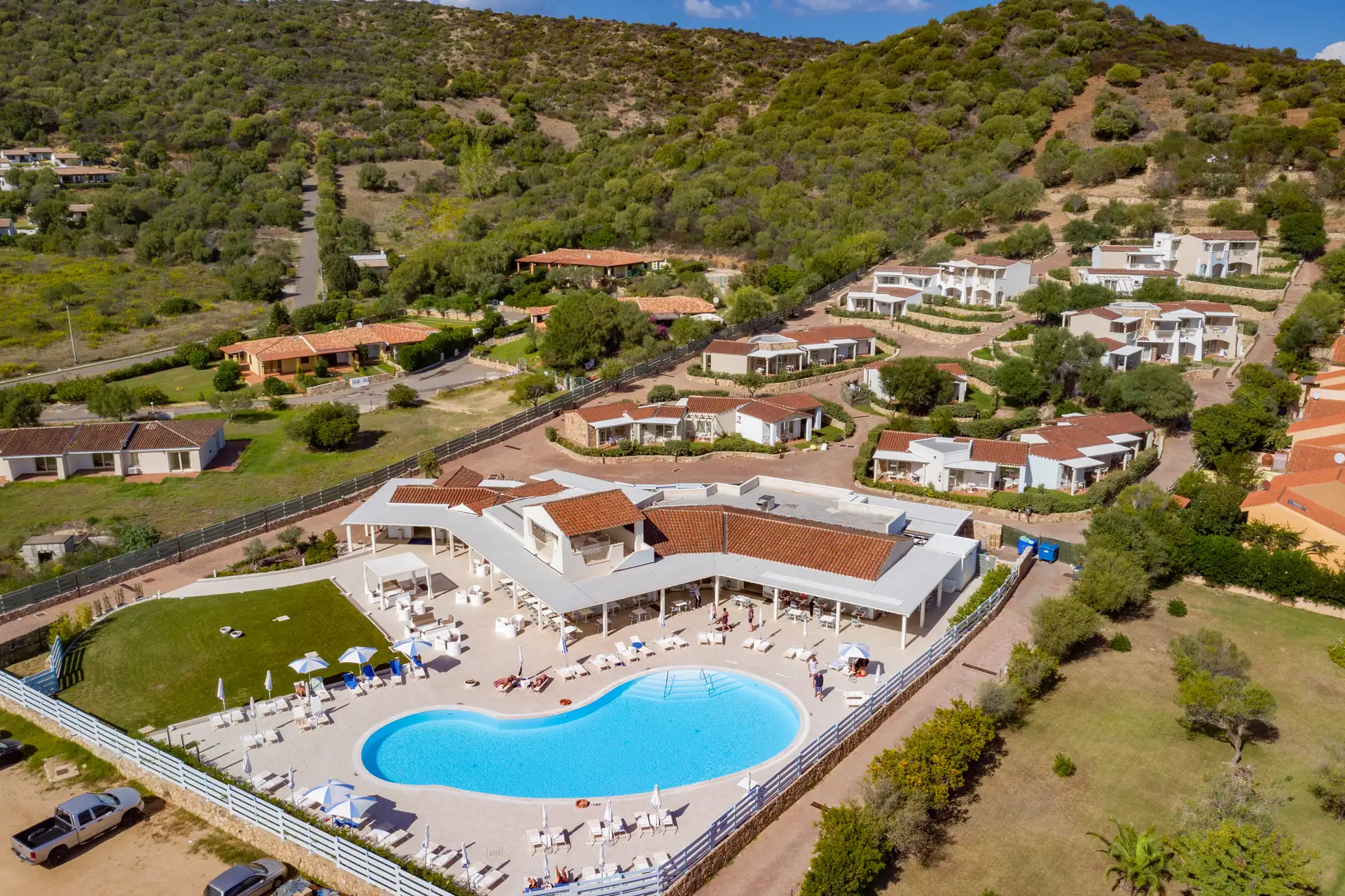 Drohnenaufnahme vom Hotel Piccolo in Porto Ottiolu mit herzförmigem Pool, Liegestühlen und Sonnenschirmen, umgeben von grünen Hügeln und mehreren weißen Gebäuden mit roten Dächern