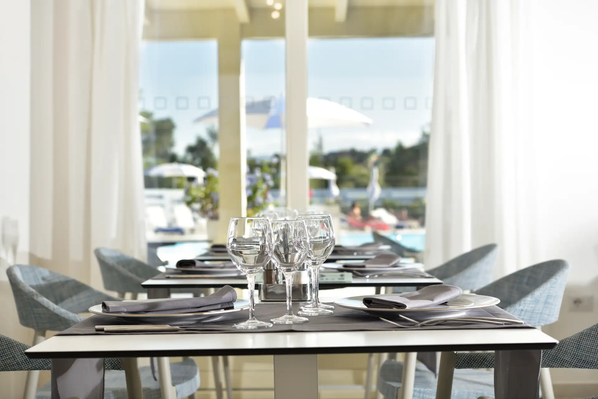 Eleganter gedeckter Tisch im Hotel Piccolo in Porto Ottiolu mit Weingläsern und Besteck in einem hellen Restaurant mit Blick auf einen Pool und Sonnenschirme