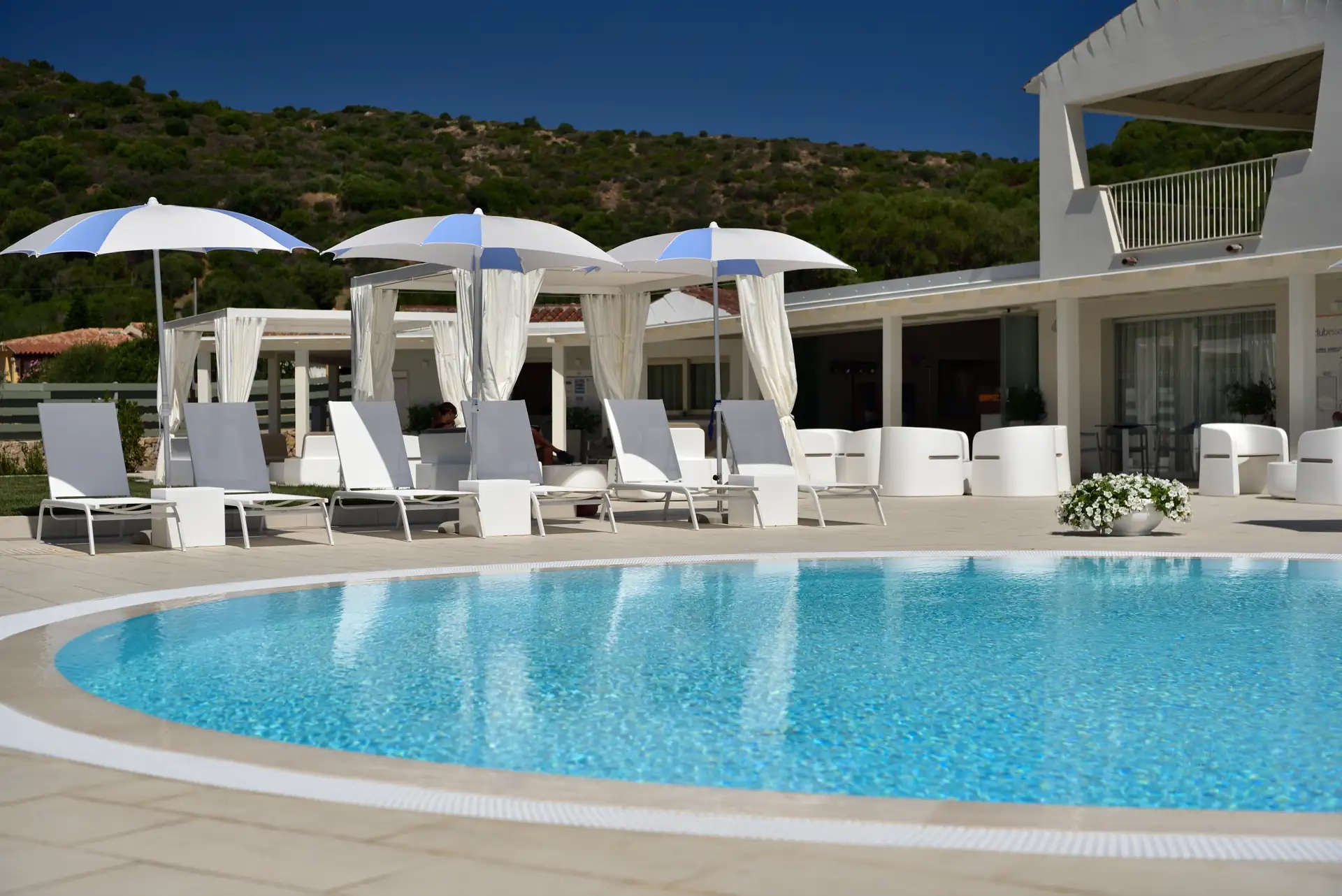Runder Pool mit klarem Wasser im Hotel Piccolo in Porto Ottiolu, umgeben von weißen Liegestühlen und Sonnenschirmen vor einem modernen Gebäude und grünen Hügeln im Hintergrund