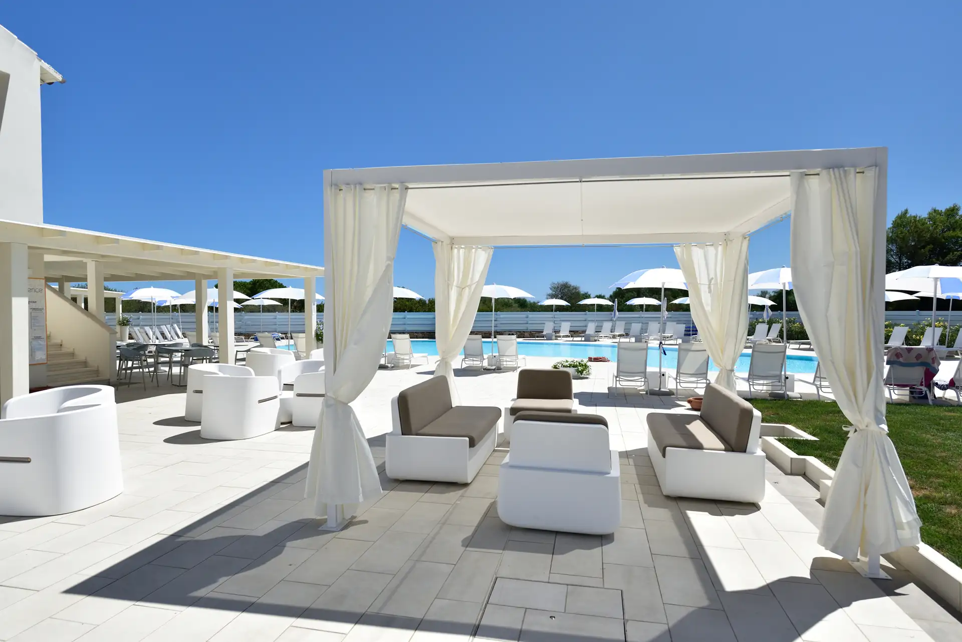 Weiße Lounge-Möbel unter einem Pavillon neben einem Pool mit Liegestühlen und Sonnenschirmen bei klarem Himmel im Hotel Piccolo in Porto Ottiolu