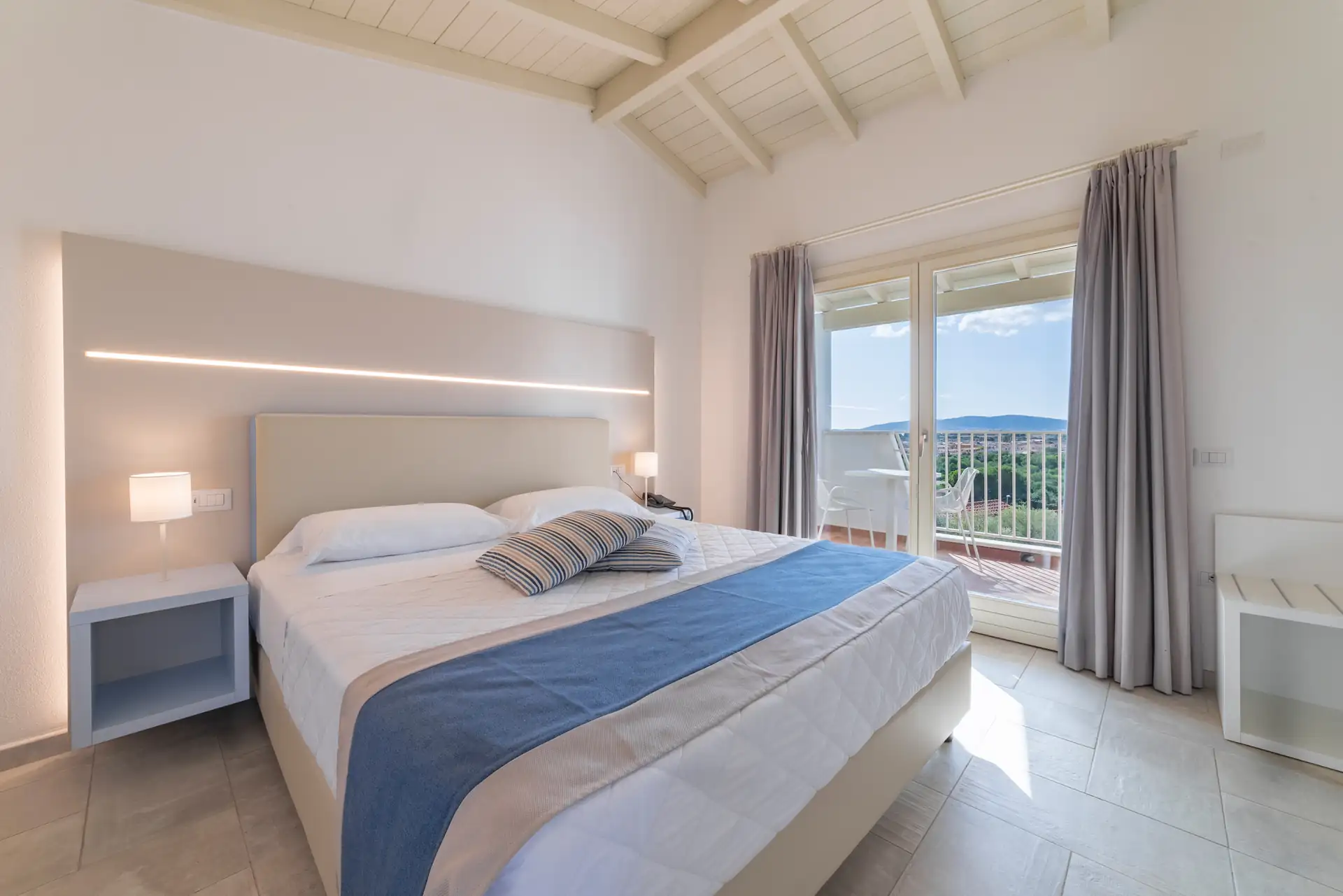 Helles Schlafzimmer im Hotel Piccolo in Porto Ottiolu mit Doppelbett, blauer und beiger Bettdecke, zwei Nachttischen mit Lampen und Balkon mit Blick ins Grüne