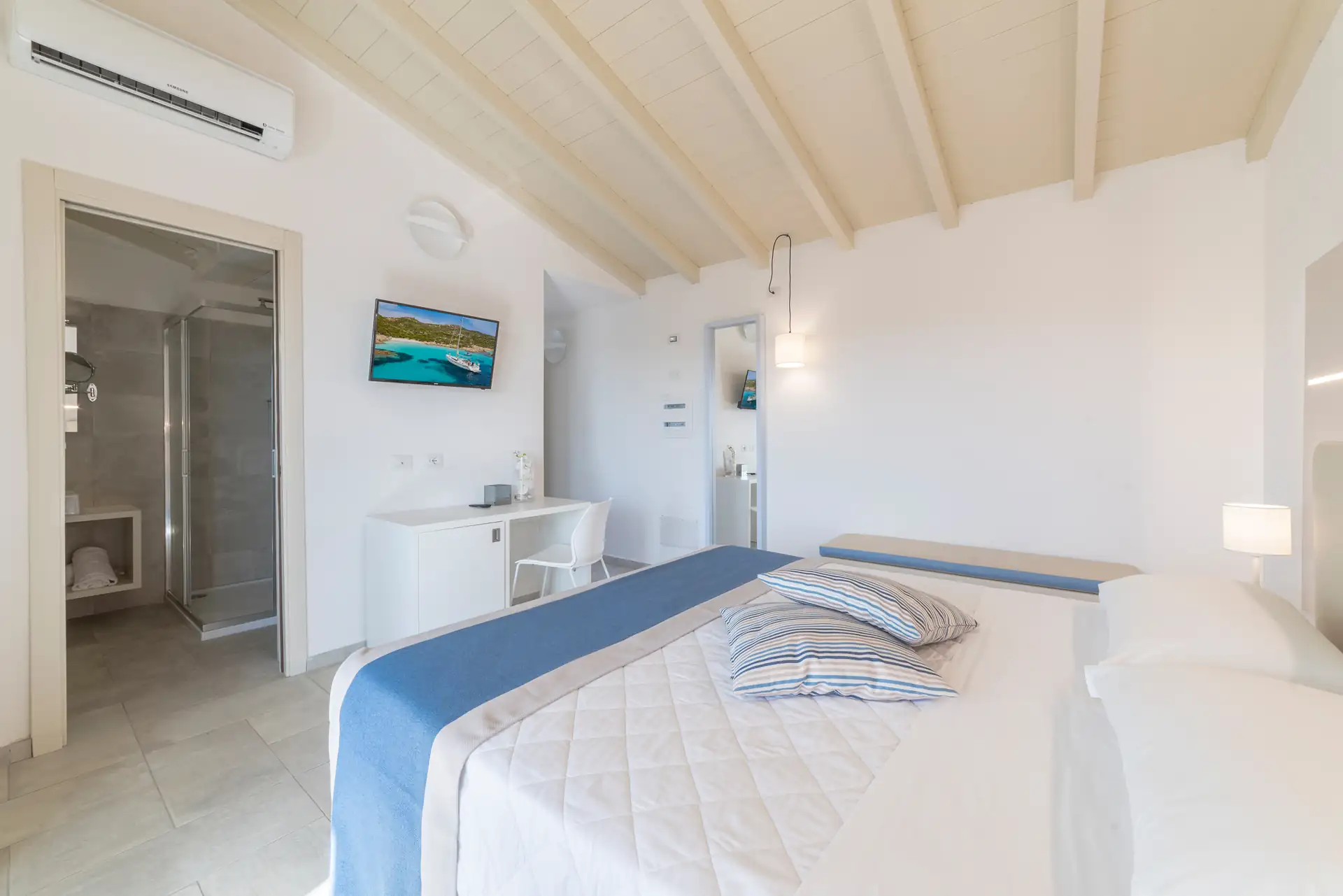 Helles Schlafzimmer im Hotel Piccolo in Porto Ottiolu mit weißem Holzbalkendecken, Doppelbett mit weißer Bettdecke und blauen Akzenten, Schreibtisch mit Stuhl, Wandfernseher und angrenzendem Badezimmer