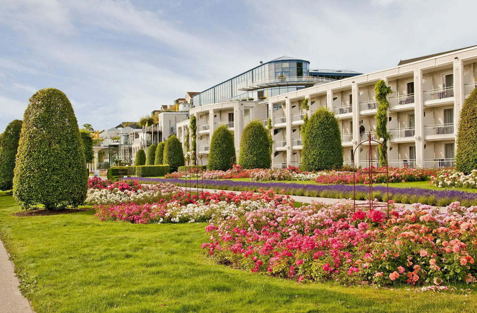 Hotelgebäude mit Balkonen neben einem gepflegten Garten mit bunten Blumenbeeten und geschnittenen Hecken unter bewölktem Himmel