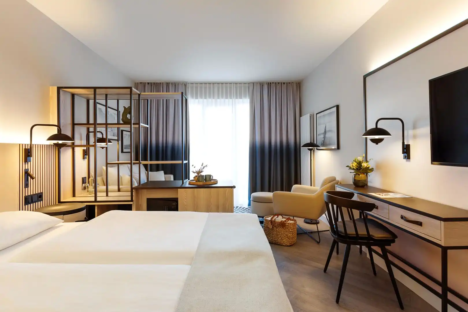 Modern eingerichtetes Doppelzimmer mit zwei Betten, Schreibtisch, Sessel, TV und Raumteiler vor Fenster mit Vorhängen