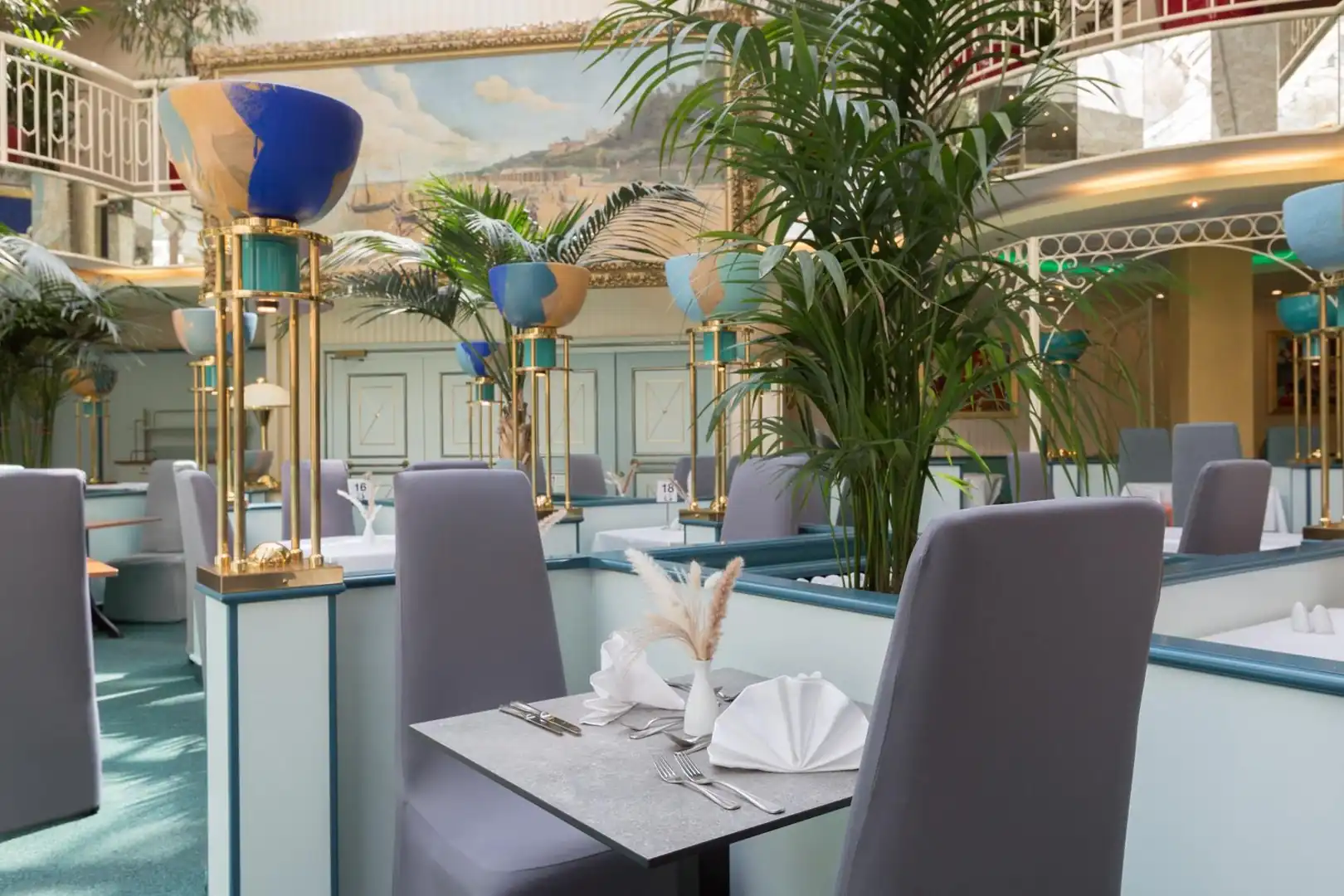 Hotelrestaurant mit gedeckten Tischen, grauen Stühlen, großen Pflanzen und Wandgemälden in hellem Ambiente