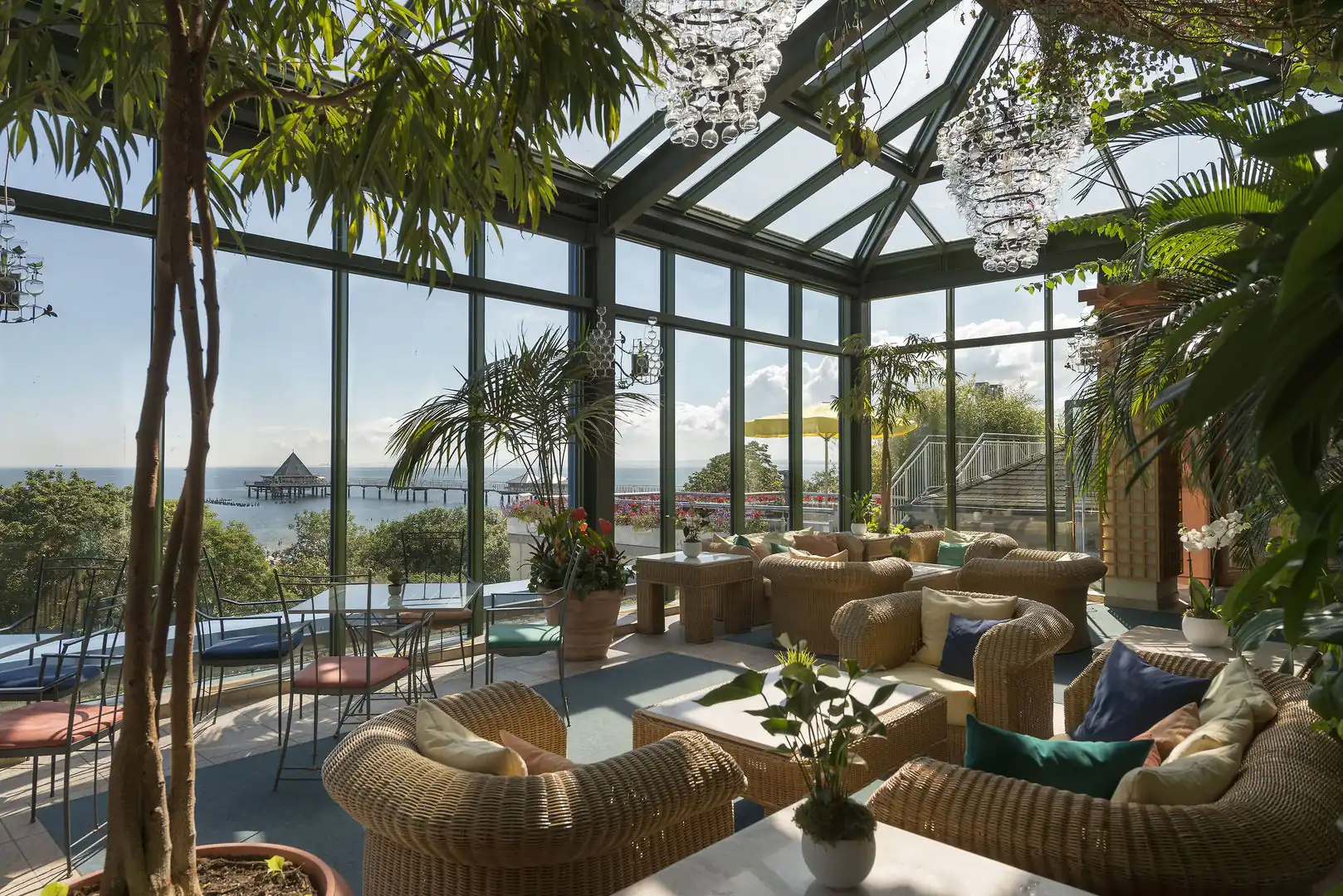 Wintergarten mit Korbsesseln, Pflanzen und Blick auf eine Seebrücke über das Meer