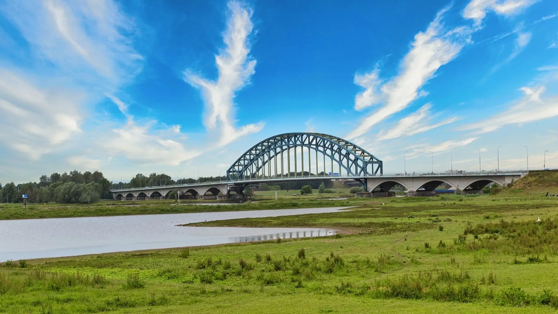 Stählerne Bogenbrücke über den Fluss IJssel bei Zwolle, umgeben von grünem Ufer und blauem Himmel mit Wolken.